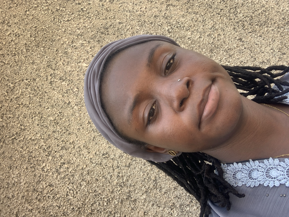 A forensic description of the scene in Bojude, Nigeria, depicts a female subject captured in a portrait-oriented, close-up photograph. The subject, a dark-skinned individual, is positioned slightly off-center to the left, facing generally forward with a neutral expression and a gaze directed slightly downwards and to the left. She wears a grey fabric headwrap covering the top of her head, with dark dreadlocks visible extending past her shoulders on both sides. A small silver stud is present in her left nostril, and a circular gold earring is visible in her right earlobe. She is dressed in a light grey garment featuring intricate white lace detailing along the neckline. A thin gold chain necklace is visible around her neck. The background consists of a uniformly textured wall, light brown or beige in color, characteristic of stucco or a rough plaster finish, evenly illuminated, suggesting natural light in an outdoor setting.
