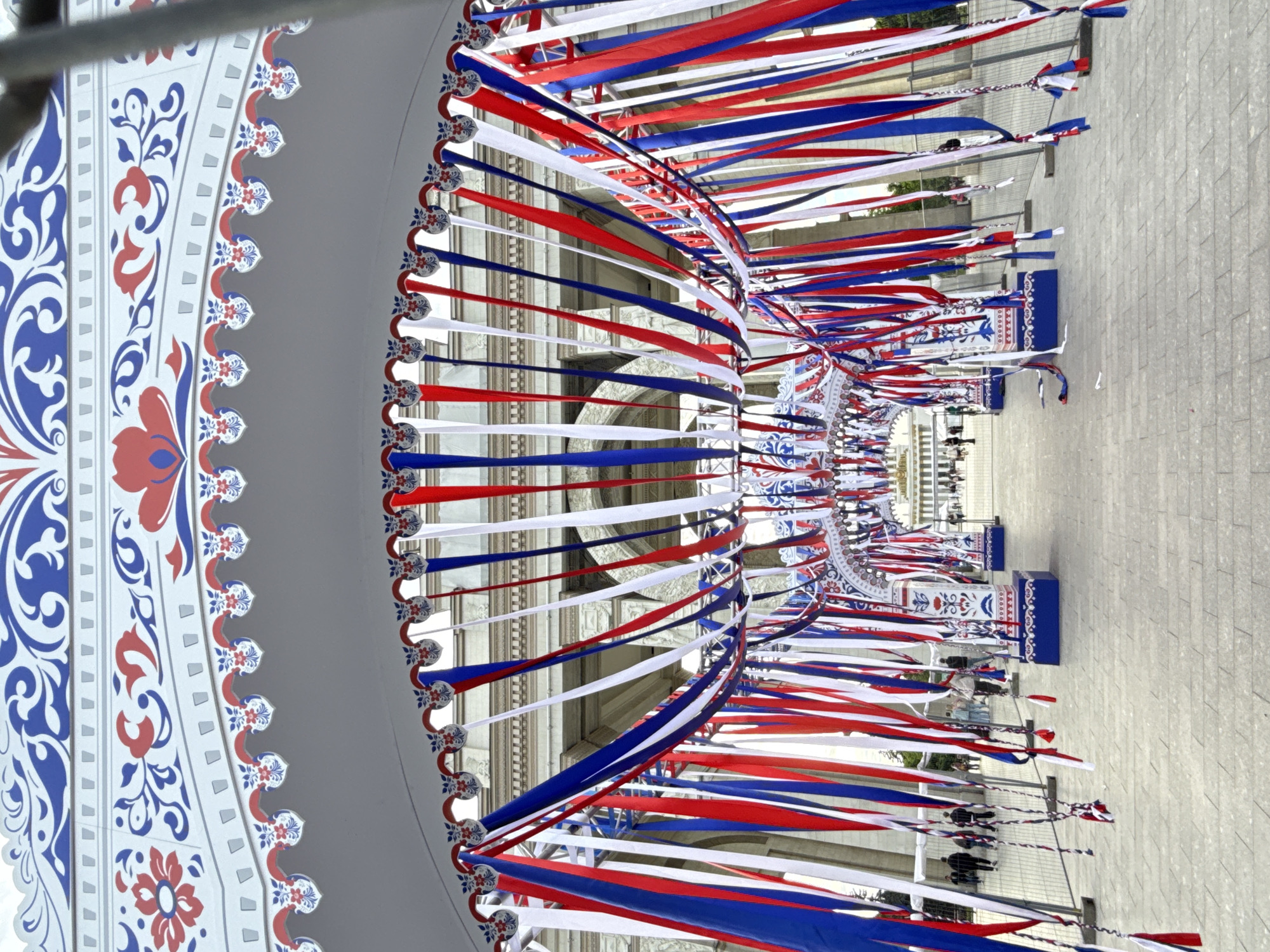 The image displays a vibrant outdoor scene in Moscow, Russia, dominated by an extensive festive decoration. A large archway or tunnel is adorned with numerous red, white, and blue ribbons, reminiscent of the Russian national flag. These ribbons hang densely, creating a dynamic canopy over a paved walkway.
The structure supporting the ribbons is elaborately decorated with traditional patterns in blue and red on a light background, featuring intricate floral and scroll motifs. Smaller, scalloped borders also exhibit similar designs, from which the individual ribbons cascade. In the background, a grand, classical-style building with arched windows and columns is visible, suggesting a significant historical or governmental landmark, consistent with Moscow's architecture.
Along the pathway beneath the ribbons, several dark blue, rectangular structures, patterned with white and red designs, are placed. These could be benches, planters, or decorative pedestals. Further down the path, several people are visible, indicating a public gathering or event.
The bright, even lighting suggests it is daytime, likely under clear weather. The overall ambiance is one of celebration and national pride, characteristic of a public holiday or special event. No discernible text is visible in the image.