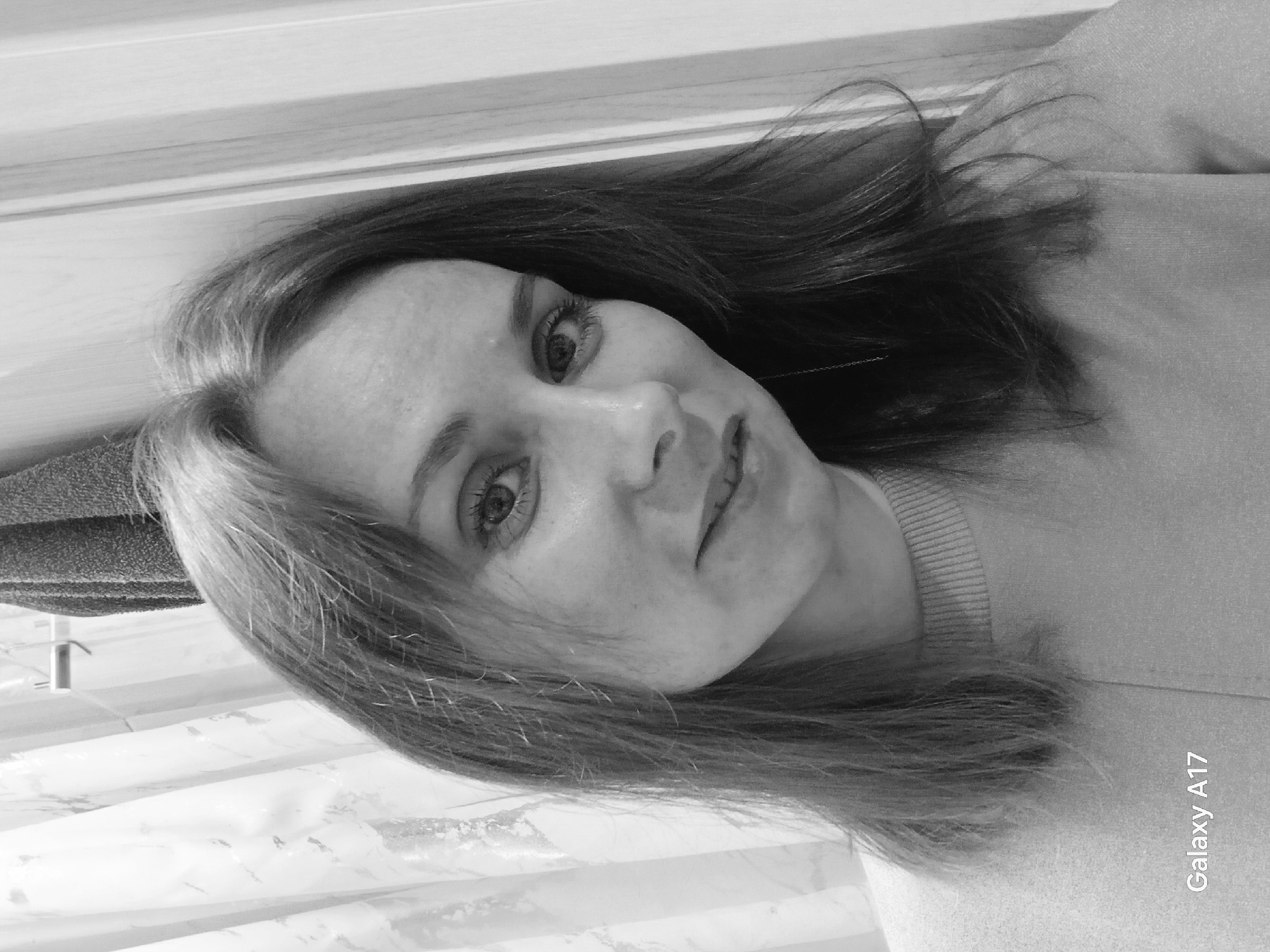 The image is a black and white selfie of a woman. She has light hair and is wearing a collared shirt. The background is blurry and indistinct. The words "Galaxy A17" are printed in the lower right corner of the image. The location cannot be determined based on the image.
