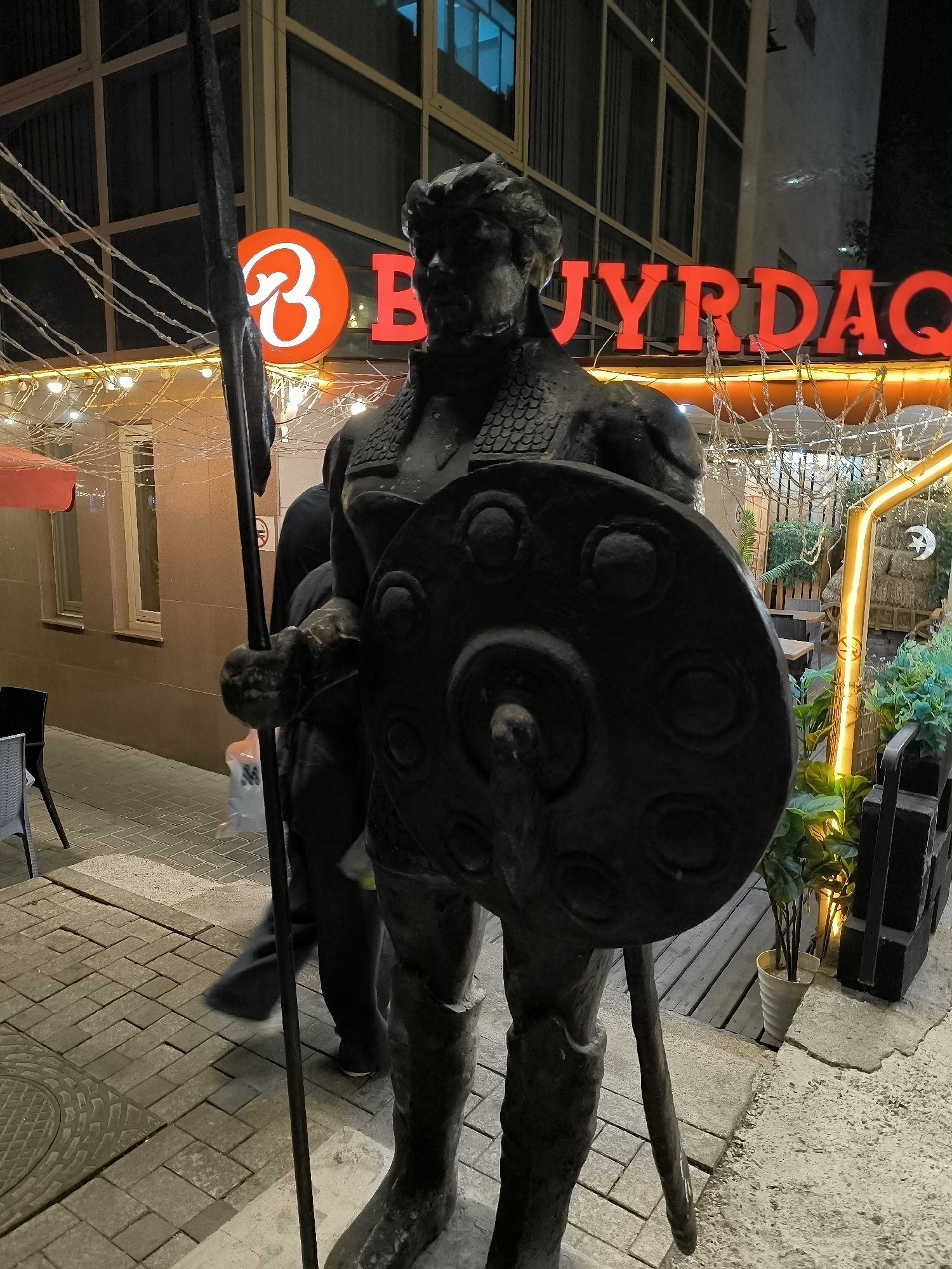 A dark bronze statue of a warrior holding a spear and shield stands prominently in front of a restaurant with a bright red neon sign reading "BUYRDAQ" in Almaty, Kazakhstan. The warrior is dressed in armor, with a segmented chest plate and what appears to be a helmet. The shield is round and decorated with circular patterns. The statue is positioned on a cobblestone sidewalk. Behind the statue, the restaurant features outdoor seating with wooden tables and chairs, plants, and string lights overhead, creating a warm ambiance. The building's facade is warm-toned, with windows and a red awning visible. The image is captured at night, with artificial lighting illuminating the scene. A person is blurred in motion to the left of the statue, suggesting movement within the frame.