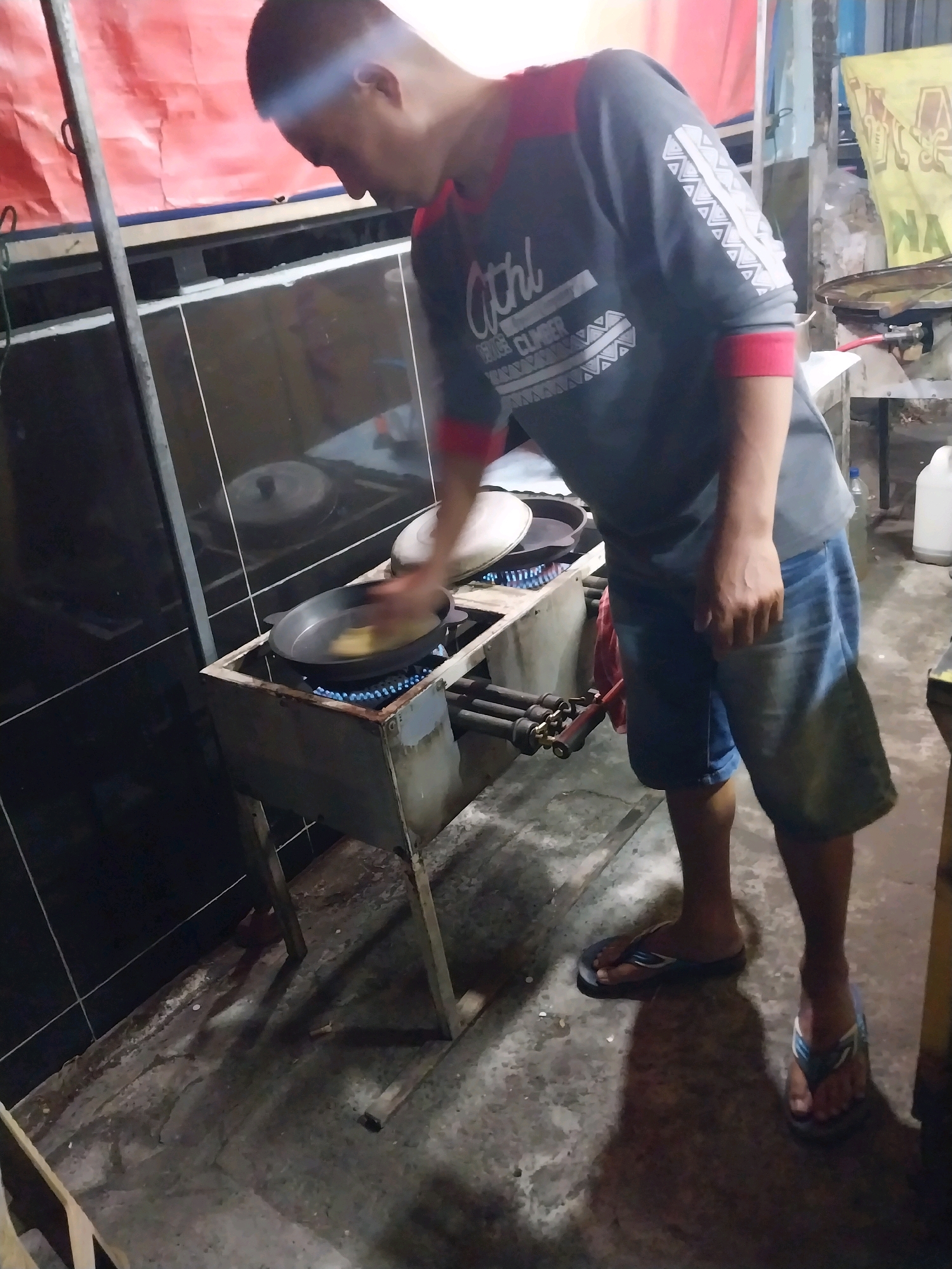 The image shows a man cooking food on a gas stove outdoors. He is wearing a gray t-shirt with red sleeves and blue denim shorts. He is also wearing flip-flops. The stove is a portable propane gas stove with two burners. The man is holding a spatula and appears to be stirring food in a pan. The background of the image shows a wall with black tiles and some colorful banners or tarps. There are also some plastic containers and other items that suggest an outdoor market or food stall setting. The scene is likely in the city of Nglegok, Indonesia, given the context provided. The ground is concrete and appears somewhat worn. The lighting suggests it is either early morning or late evening.