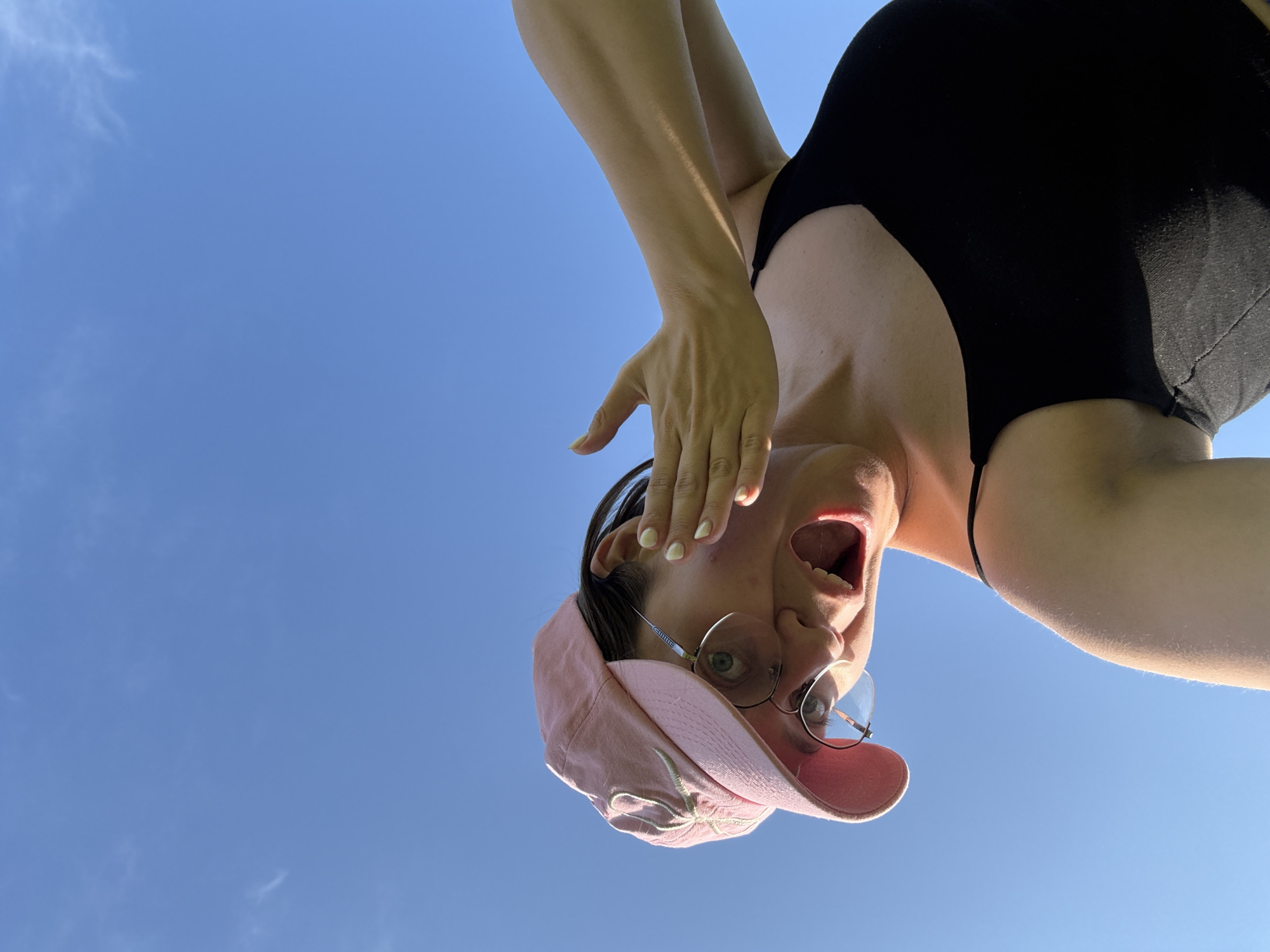 The image shows a woman with fair skin and dark hair wearing a pink baseball cap and a black tank top. She is wearing round glasses and has her mouth open, appearing surprised or yelling. Her right hand is raised, partially covering her face. The background is a clear blue sky with a few wispy clouds. The woman is positioned in the foreground, looking upwards at the camera, which is angled from below. The lighting suggests it is daytime. The image does not provide any information about a specific location in Dokshitsy, Russia.