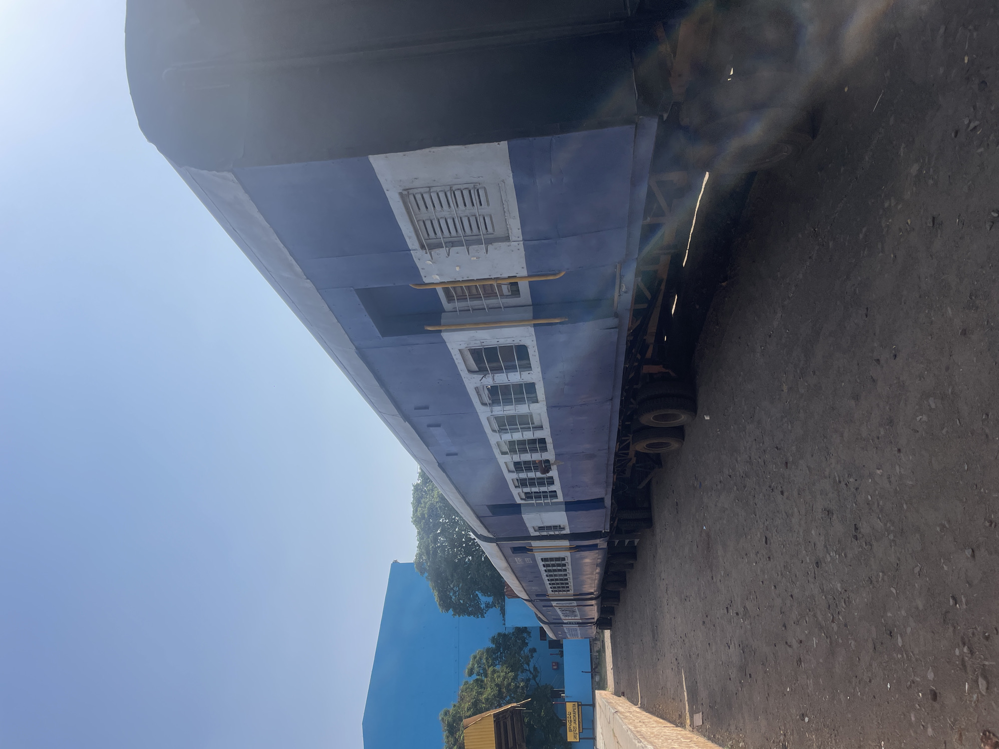 The image shows a blue and white train car at the Anajpur train station in Anajpur, India. The train is parked on a dirt surface, with a blue building and trees visible in the background.