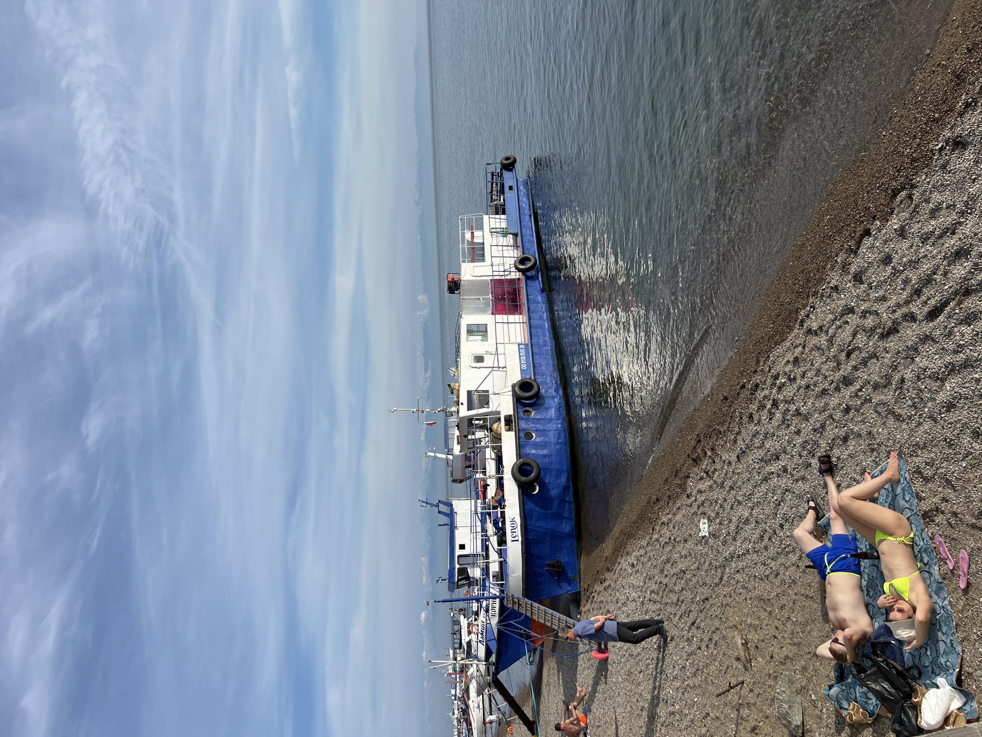In Listvyanka, Russia, a tranquil scene unfolds along the pebbled shore of Lake Baikal. The sun casts a gentle light on a calm body of water where several boats are docked. In the foreground, two people are relaxing on a blanket. A man in blue swimming trunks lies on his back with his hands behind his head, while a woman in a yellow bikini reclines beside him. A bottle lies on the blanket between them, and pink flip-flops are scattered nearby. Behind them, a man in a dark blue shirt and black pants stands on the shore, looking towards the boats. Further back, a large blue and white boat is moored, with a ladder leading from its deck to the shore. The boat has "Lenok" written on its side in blue letters. Other boats are visible in the background, suggesting a small harbor. The sky above is a pale blue, adorned with wispy white clouds. In the distance, a hazy mountain range can be seen along the horizon. The overall atmosphere is one of peaceful leisure.