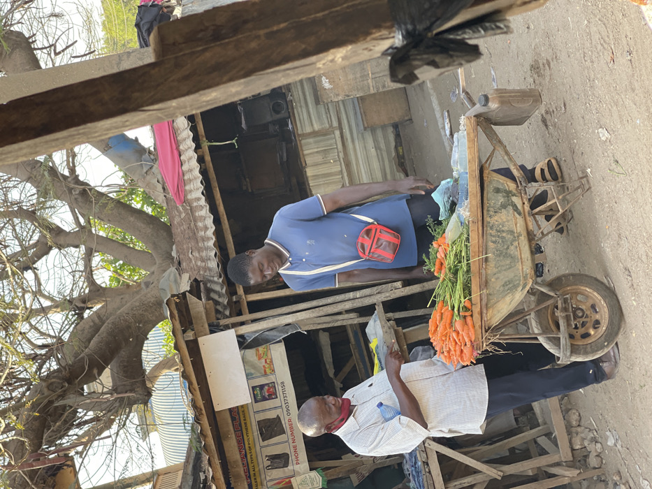 Two men are standing in a street market. One man is wearing a blue shirt and a red crossbody bag, standing near a wheelbarrow filled with carrots. The other man is wearing a white shirt and holding a piece of paper. They are positioned in front of a small wooden structure with a corrugated metal roof. Trees and various items are visible in the background. The photograph appears to be taken in a low-income or developing area.