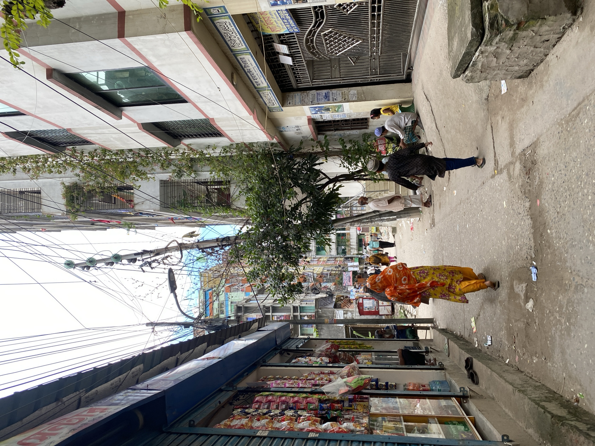 This is a ground-level, eye-level shot looking down a narrow alleyway in Dhaka, Bangladesh. The alley is lined with multi-story buildings on both sides. On the left, there is a shop with its shutters partially open, displaying various packaged goods like snacks and drinks. The shop has a blue awning and a metal grate. Further down the alley, more buildings with colorful facades and balconies are visible.
On the right side of the alley, a white building with red trim is prominent. It has several windows, some with metal grills, and is covered in vines. A large metal gate is visible in front of this building, along with a sign in Bengali script. There are also several people in the alleyway.
In the foreground, a woman wearing a vibrant orange and yellow floral dress walks away from the camera. Further down the alley, a man in a black patterned outfit and blue pants walks towards the background. Several other individuals, including men and a child, are seen sitting and standing on the right side of the alley.
The alley itself is paved with concrete and shows signs of wear and tear. There are scattered pieces of trash and debris on the ground. Overhead, a complex network of electrical wires and utility poles crisscrosses the sky. The overall impression is of a busy, densely populated urban environment.