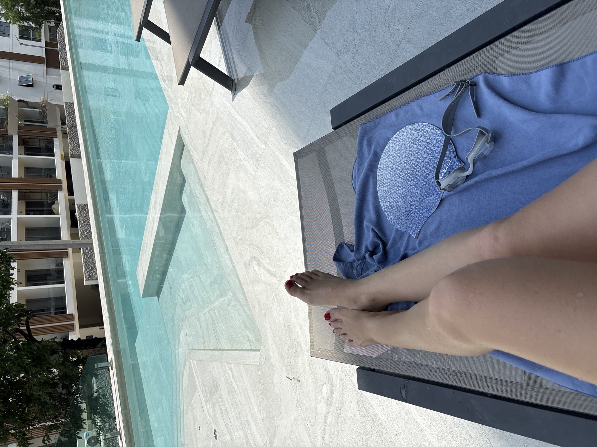A poolside scene in Choeng Thale, Thailand. A lounge chair is visible, draped with a blue towel upon which rests a blue patterned swimming cap and grey goggles. A fair-skinned person's legs extend from the chair, feet adorned with red nail polish. Adjacent to the chair is a swimming pool with turquoise water, flanked by a tiled patio area. In the background, a multi-story building is observed, with trees partially obscuring its lower levels.