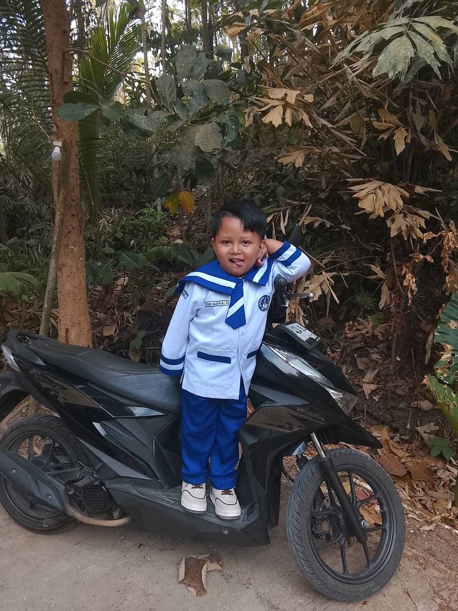A young child wearing a white and blue school uniform stands on a black scooter in Kertahayu, Indonesia. The child has dark hair and is smiling at the camera, with their right hand touching their head. The scooter has a black body and black wheels, with the "HONDA" logo visible on the front. The background is a dense forest with trees, foliage, and fallen leaves. The ground is dirt.