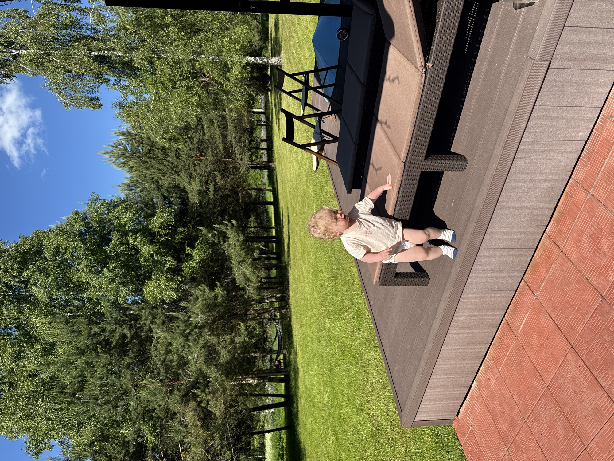 This image captures an outdoor scene in Копнино, Russia, under bright sunlight. In the immediate foreground on the left, a section of red, square-patterned paving tiles is visible. Adjacent to this, occupying the lower half of the frame and extending to the right, is a raised platform constructed from dark brown composite or wooden decking material.
On this platform, a young child with curly blonde hair is positioned towards the right side of the frame, facing left. The child is wearing a light-colored t-shirt, a white diaper, and blue and white striped socks, and is standing upright with their right hand resting on a large, brown woven outdoor lounge chair equipped with brown cushions. Further back on the platform, two dark brown wooden folding chairs are present, alongside a blue tarp or cover.
The background consists of a vast, meticulously manicured green lawn. Beyond the lawn, a dense forest line features a mix of tall conifer trees and deciduous trees with vibrant green foliage. In the far left background, a hammock is suspended between two trees, with a person partially discernible resting within it. The sky is clear and blue, accented by a few scattered white cumulus clouds. Strong shadows are cast across the decking and lawn, indicating direct, intense sunlight.