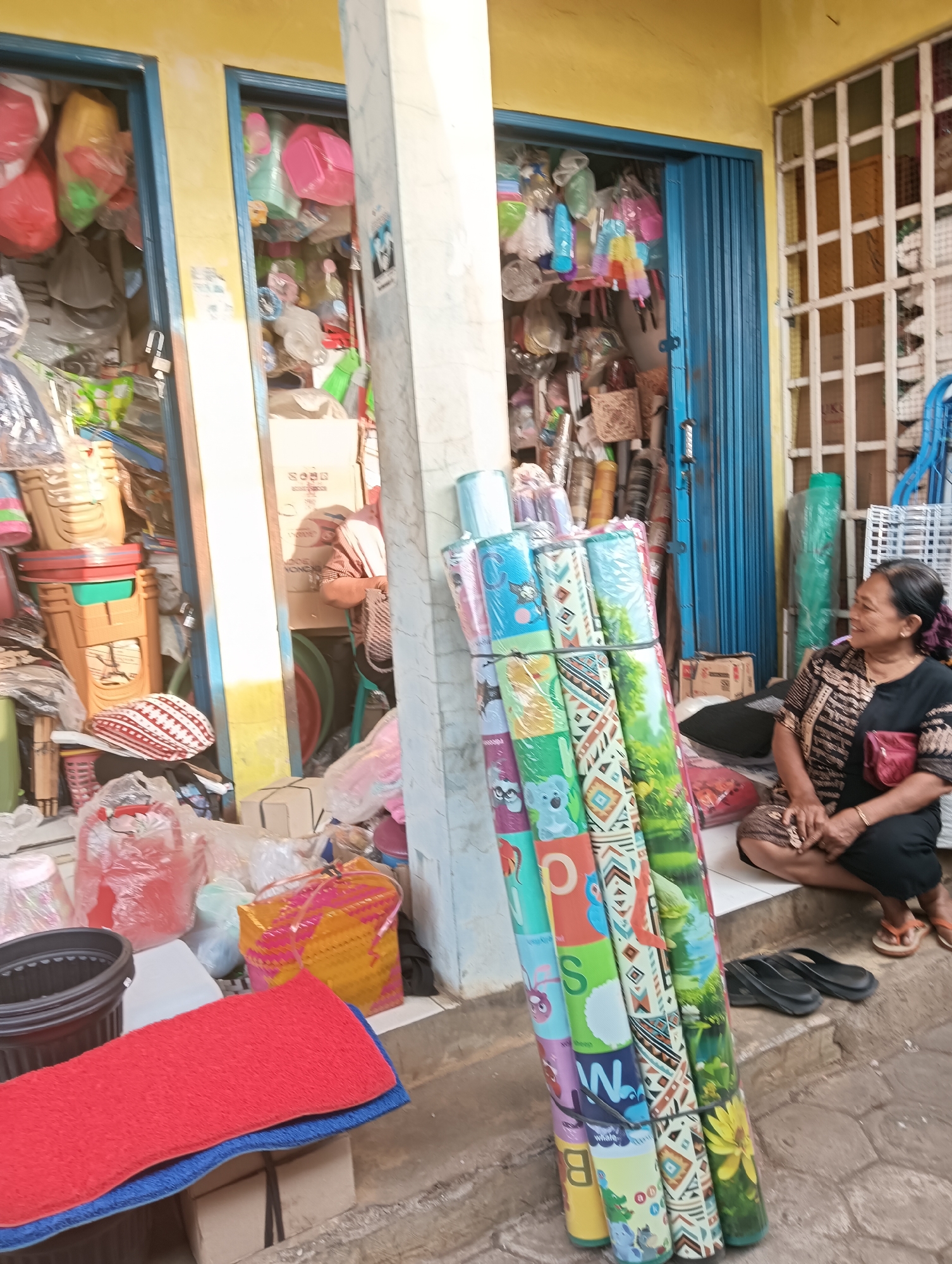 In front of a shop in Tayu, Indonesia, a woman sits on steps next to rolled-up mats. The shop's facade is painted yellow, and its entrances are covered by blue accordion doors. The shop appears to be a retail establishment selling various goods, including plastic containers, bags, and woven baskets, which are visible through the open doorways and piled outside. The woman, wearing a dark patterned top and black pants, has a small red pouch attached to her belt. She is looking towards the left side of the frame. Several rolled-up mats, adorned with colorful patterns and images of animals and nature, are stacked vertically to the left of the woman. The ground in front of the shop is paved with cobblestones. To the right of the shop, a window is covered by a white metal grate. The overall scene depicts a bustling market or street-side shop environment.