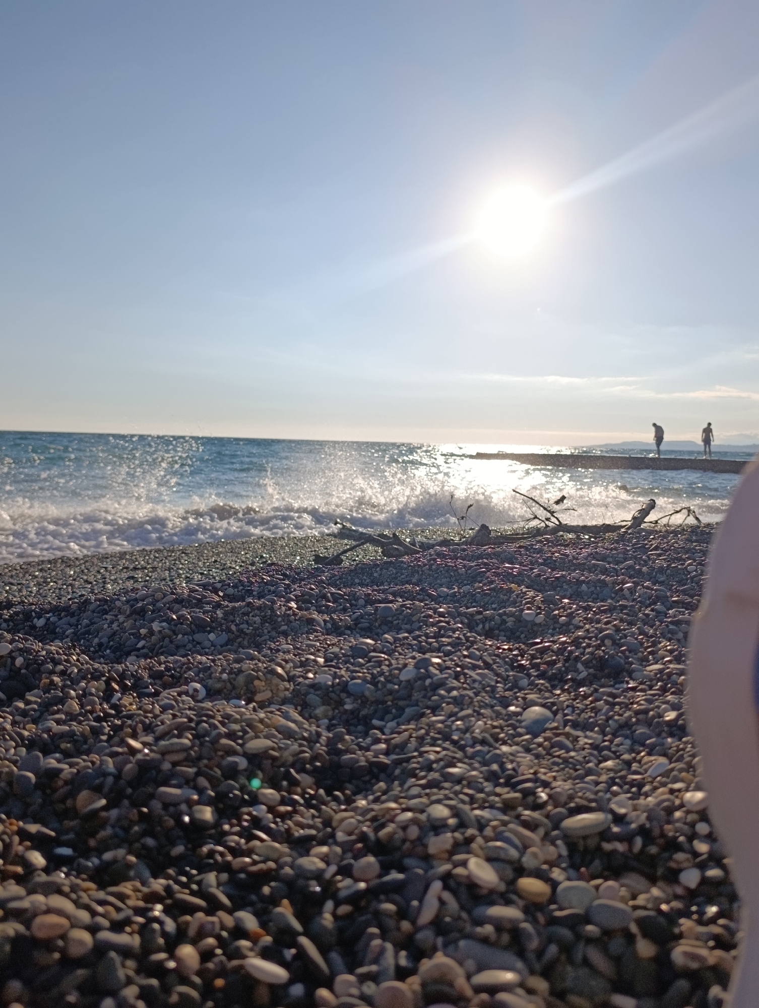 The image depicts a sunny day at a pebble beach in Eshyra, Abkhazia. The sun, positioned in the upper center, is bright and casting a strong glare, with a light beam extending diagonally across the sky. The sea is a vibrant blue with white-crested waves crashing onto the shore. The foreground is dominated by a dense covering of various sized pebbles. In the mid-ground, two figures are visible standing on a pier that extends into the sea. A broken branch lies on the pebble shore near the water's edge. A blurred object, possibly a person's shoulder or arm, is in the extreme lower right corner of the frame.