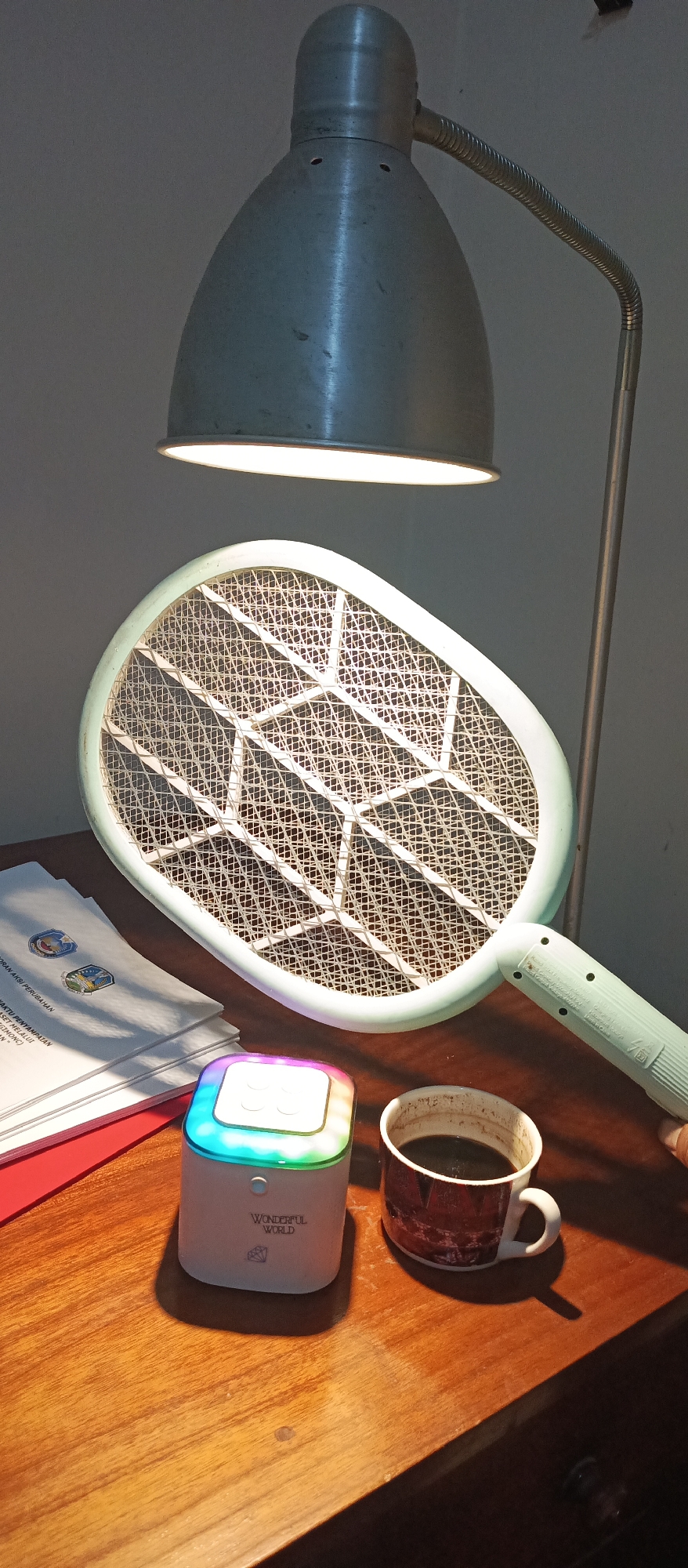 The image shows a desk in Jatisari, Indonesia. On the desk are a lamp, a mosquito racket, a stack of papers, a small colorful device, and a mug of coffee. The lamp is metallic and angled towards the mosquito racket. The mosquito racket is light green and has a mesh surface. The stack of papers has some text and logos on it. The small device is white and glows with rainbow colors. The mug is white with a red and black pattern and is filled with dark liquid, presumably coffee. The desk appears to be made of wood. The lighting suggests it is nighttime or a dimly lit room.