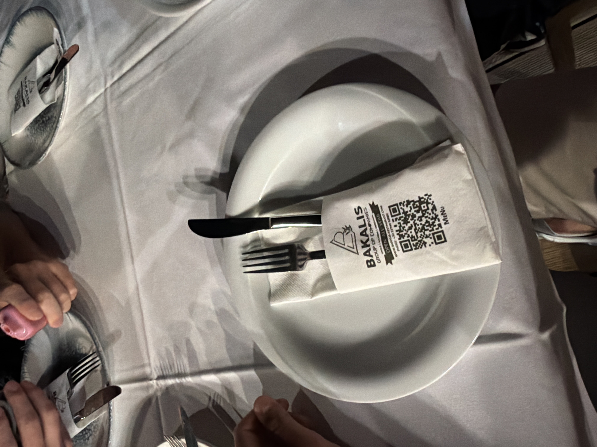 Here is a detailed description of the image:
The image shows a table setting in what appears to be a restaurant, featuring white tablecloths, plates, cutlery, and napkins. Two plates are fully visible, with a third only partially visible in the lower left corner. Each plate has a knife and fork set wrapped in a white napkin. The napkin on the main plate has the logo "BAKALIS GROUP OF COMPANIES" printed on it, along with a QR code and contact information. Several people's hands are visible around the plates, suggesting that people are seated at the table. The lighting appears to be indoor and somewhat soft, casting shadows on the tablecloth and plates. The presence of the "BAKALIS" logo and a phone number with the +30 country code suggest the location is in Greece, specifically within the city of Pallini Municipal Unit, where Bakalis restaurant exists.