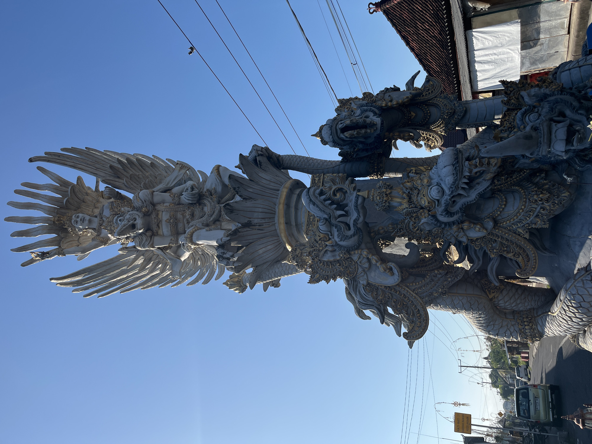 A large, ornate statue depicting Garuda, a mythical bird-like creature from Hindu mythology, dominates the frame, set against a clear blue sky. The statue is a complex sculpture, with Garuda at the apex, perched on a lotus-like base, flanked by a multi-headed dragon figure. The dragon is detailed with intricate carvings and gold accents, and its mouths are open, revealing sharp teeth. The statue is positioned near a road in Sukawati, Indonesia, with power lines and utility poles visible in the background. On the left side of the road, a yellow sign with Indonesian text is visible, and several vehicles, including a light-colored SUV, are on the road. Traditional Balinese architecture, with tiled roofs, is visible to the right of the statue. The scene appears to be a public space, possibly a roadside monument or landmark in a bustling town.