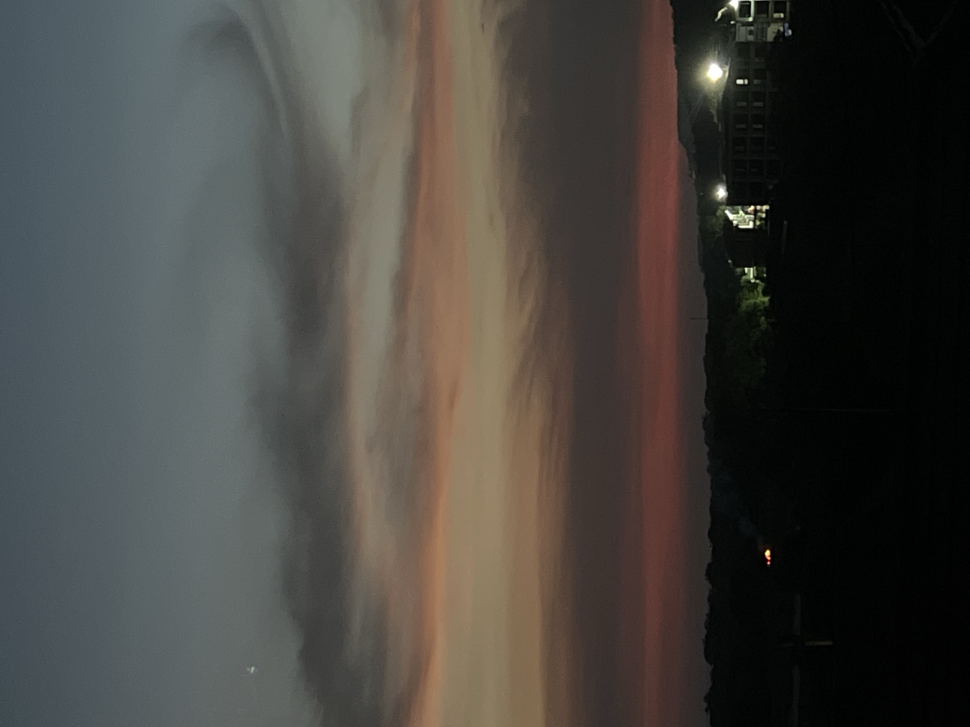 The image captures a dramatic twilight sky, likely a sunset, over a distant landscape featuring buildings. The sky, which occupies most of the frame, displays a striking gradient of colors. Starting from a deep blue-grey in the upper left, it transitions through soft whites, light oranges, and pinks, culminating in a rich, reddish-orange hue along the horizon line on the right. Wispy, swirling cloud formations are visible, particularly in the cooler, bluer sections of the sky, adding texture and depth to the celestial view.
On the far right, silhouetted against the colorful sky, is a dark, indistinct landscape or distant urban area. Within this dark silhouette, several multi-story buildings are visible. These structures are punctuated by artificial lights, which glow brightly against the deepening darkness. Notable lights include a prominent bright white-yellow illumination from a building and several smaller, warmer-toned lights scattered across the distant landmass, suggesting ongoing activity in the area as night approaches.
The scene depicts the transition between day and night, characterized by the vibrant sky colors and the activation of artificial lights. No people are visible, and there is no discernible text in the image. The overall impression is one of serene atmospheric beauty.