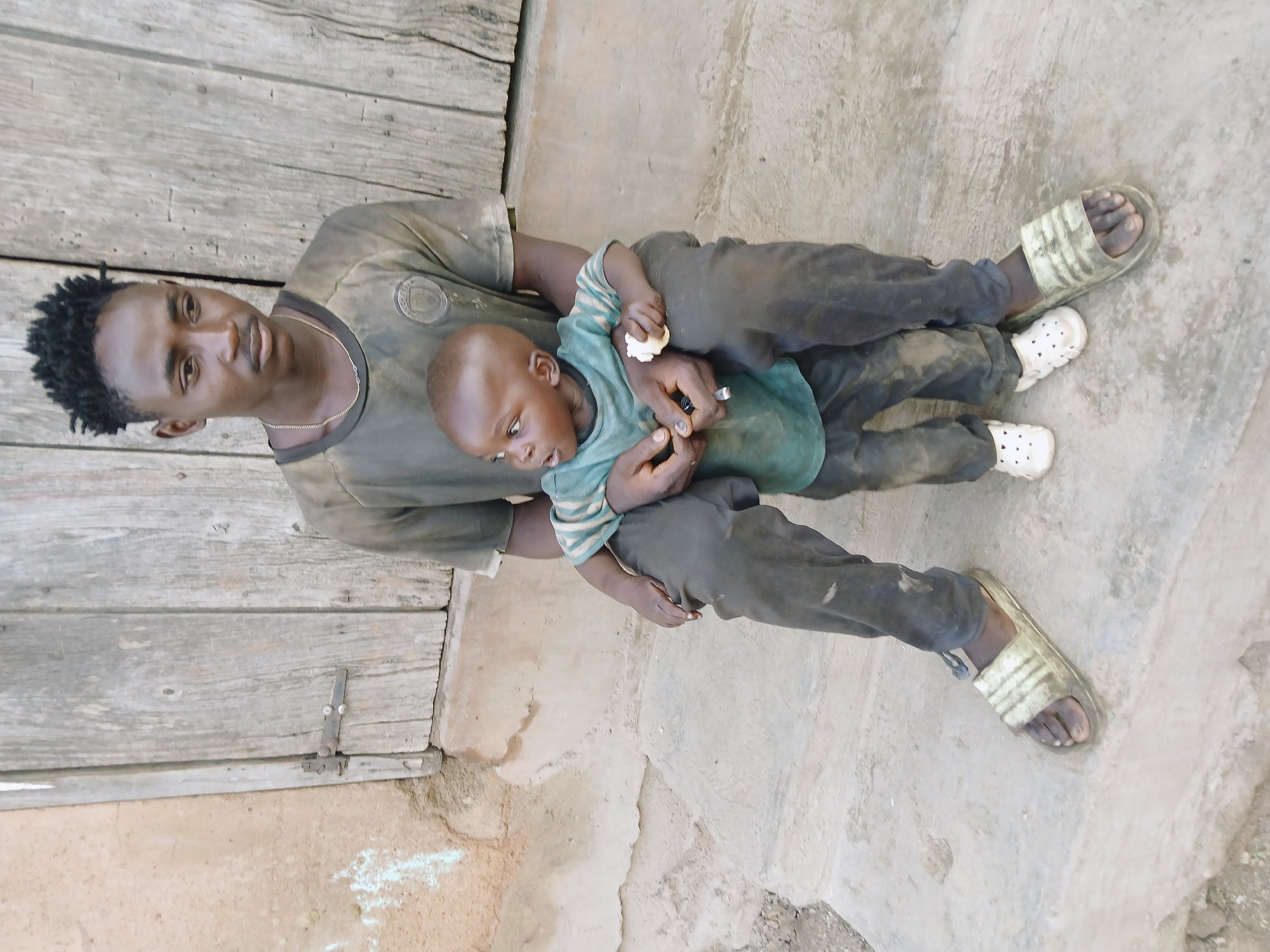 An outdoor, portrait-oriented image, rotated 90 degrees counter-clockwise, depicts an adult male holding a young child.
The adult, positioned on the left side of the frame, has dark skin and short, dark, textured hair, possibly dreadlocks or braids. He wears a dark, possibly olive-green, short-sleeved t-shirt with some subtle patterning or logo on the chest, and dark trousers. His feet, clad in light-colored, striped slide sandals, are visible towards the lower right of the image. He looks directly towards the camera with a neutral to slightly contemplative expression. A thin, light-colored chain is visible around his neck.
The child, held in the adult's lap or arms, also has dark skin and a closely shaved head. They are dressed in a light green t-shirt with darker green or blue stripes, and dark trousers. Small white, possibly foam or rubber, sandals are on the child's feet. The child is holding a small, light-colored, irregular object (resembling a piece of bread or a snack) in their right hand, and a small, dark, cylindrical object in their left hand, looking down intently at what they are holding.
Behind the adult and child, particularly to the left, is a weathered wooden wall or door constructed from horizontal planks. A simple metal latch is visible on this wooden surface. The ground beneath them is a light grey, uneven concrete or compacted dirt surface, showing textures and minor discolorations. The lighting suggests it is daytime. There is no visible text in the image. The scene conveys a casual, everyday moment between an adult and child in what appears to be a modest outdoor setting.