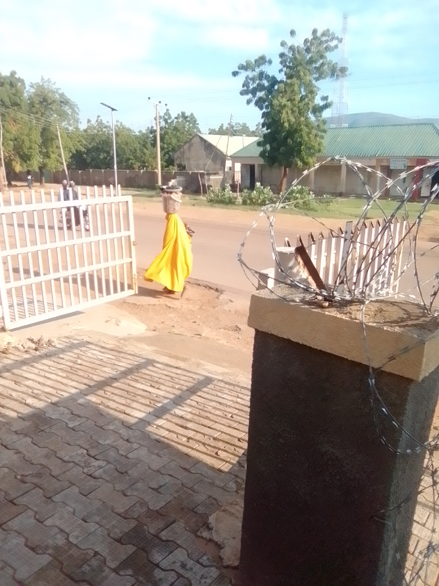 A person wearing a bright yellow garment and carrying a basket on their head walks past a white metal gate in Galadima, Nigeria. The person is walking on a dirt path next to a paved area with a geometric brick pattern. In the background, there are trees, buildings, and a cell tower. A barbed wire fence is visible in the foreground, wrapped around a concrete pillar. The sky is clear and blue.