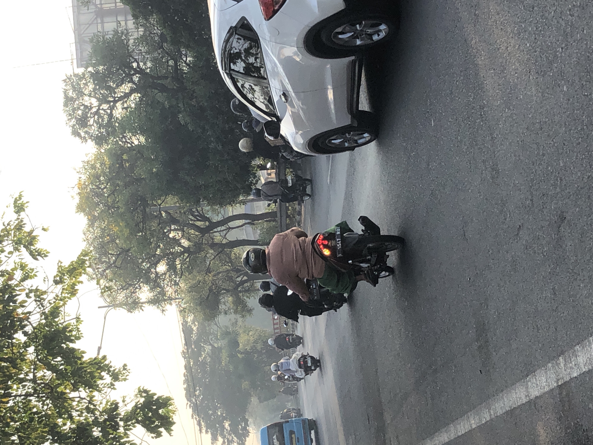 In Kludan, Indonesia, a motorcycle rider wearing a helmet and a light brown long-sleeved shirt is seen from behind, riding a motorcycle with a green cloth draped over the back. The motorcycle's license plate is visible and reads "L 5547 RQ". The rider is on a paved road with a white lane marker to their left, traveling alongside other motorcycles and a white SUV. The environment suggests a street scene with trees lining the road and a hazy, possibly early morning or late afternoon, sky.