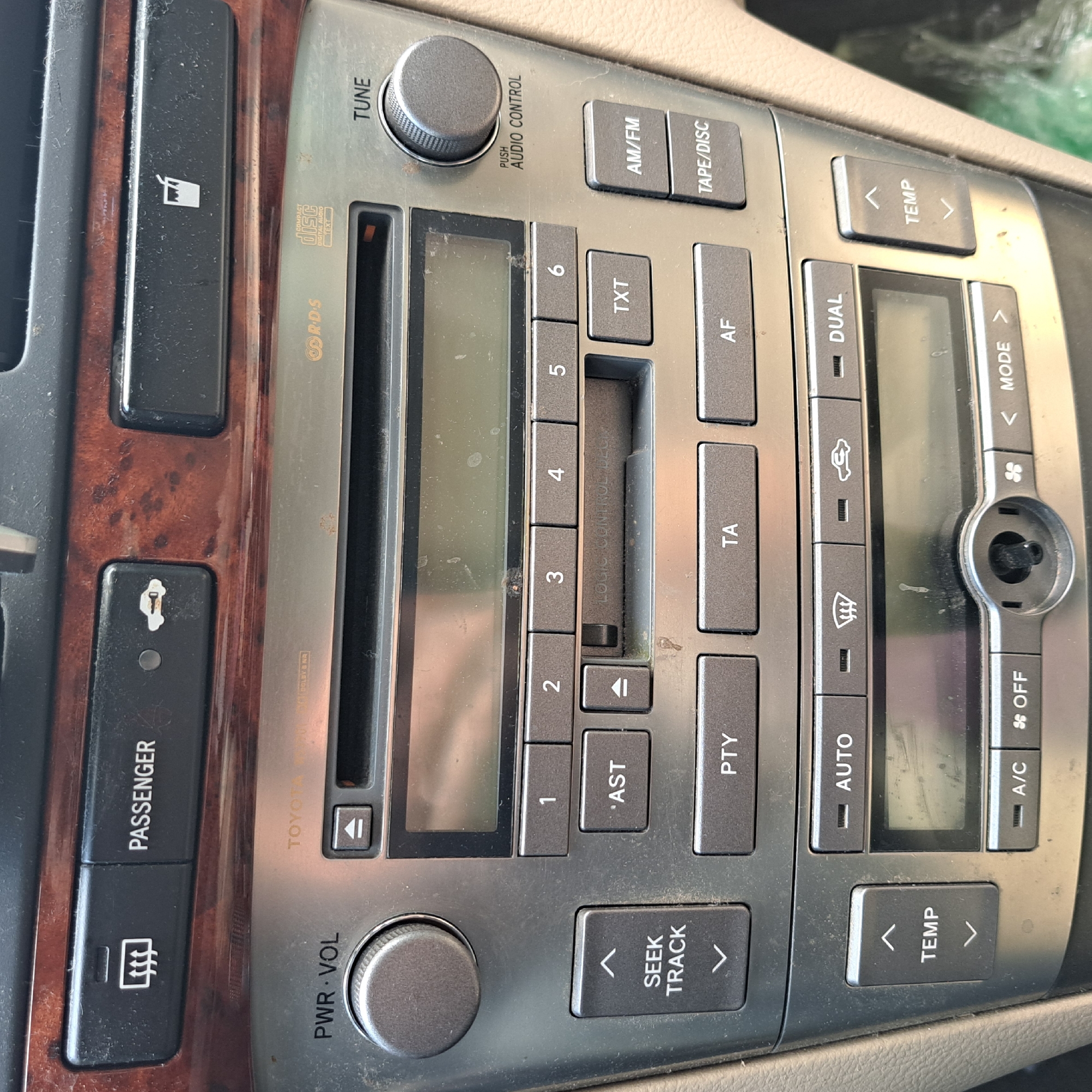 The image shows a close-up of a car's center console. The console features a car stereo with a display screen, buttons for radio tuning, track selection, and audio control. There are also climate control buttons with icons for defogging and air circulation, as well as temperature adjustment buttons. A power button for the passenger side is visible at the top left, along with a button to control the fuel door. The car's interior trim appears to be wood-grain. The image is undated and does not provide any information about its location in Odesa, Ukraine, or the presence of any people.