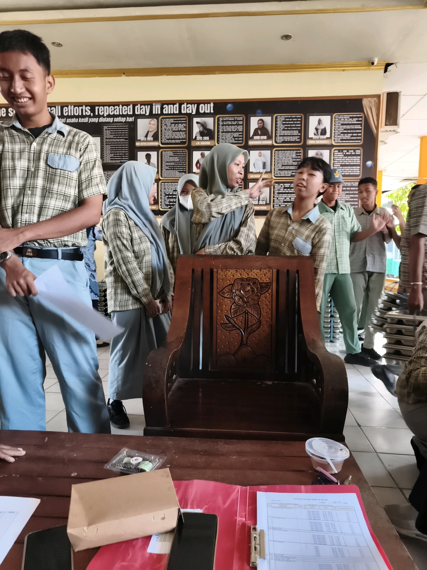 The image shows a group of students, likely in a school setting, gathered around a wooden chair. The environment appears to be indoors, possibly a classroom or common area, with a bulletin board featuring framed photographs and text in the background. Several students are wearing uniforms consisting of plaid shirts and light blue pants or skirts. One student in the foreground, wearing a grey hijab and plaid shirt, is gesturing towards another student, a boy also in a plaid shirt, who is looking upwards. Other students are standing around them. On a wooden table in the foreground, there are some documents, a brown paper bag, a clear plastic container with food, and a mobile phone. The overall scene suggests a casual interaction or possibly a presentation among students. Based on the text visible on the bulletin board, which appears to be in Indonesian, and given that the request specifies Makassar, Indonesia, the location is presumed to be a school within that city.