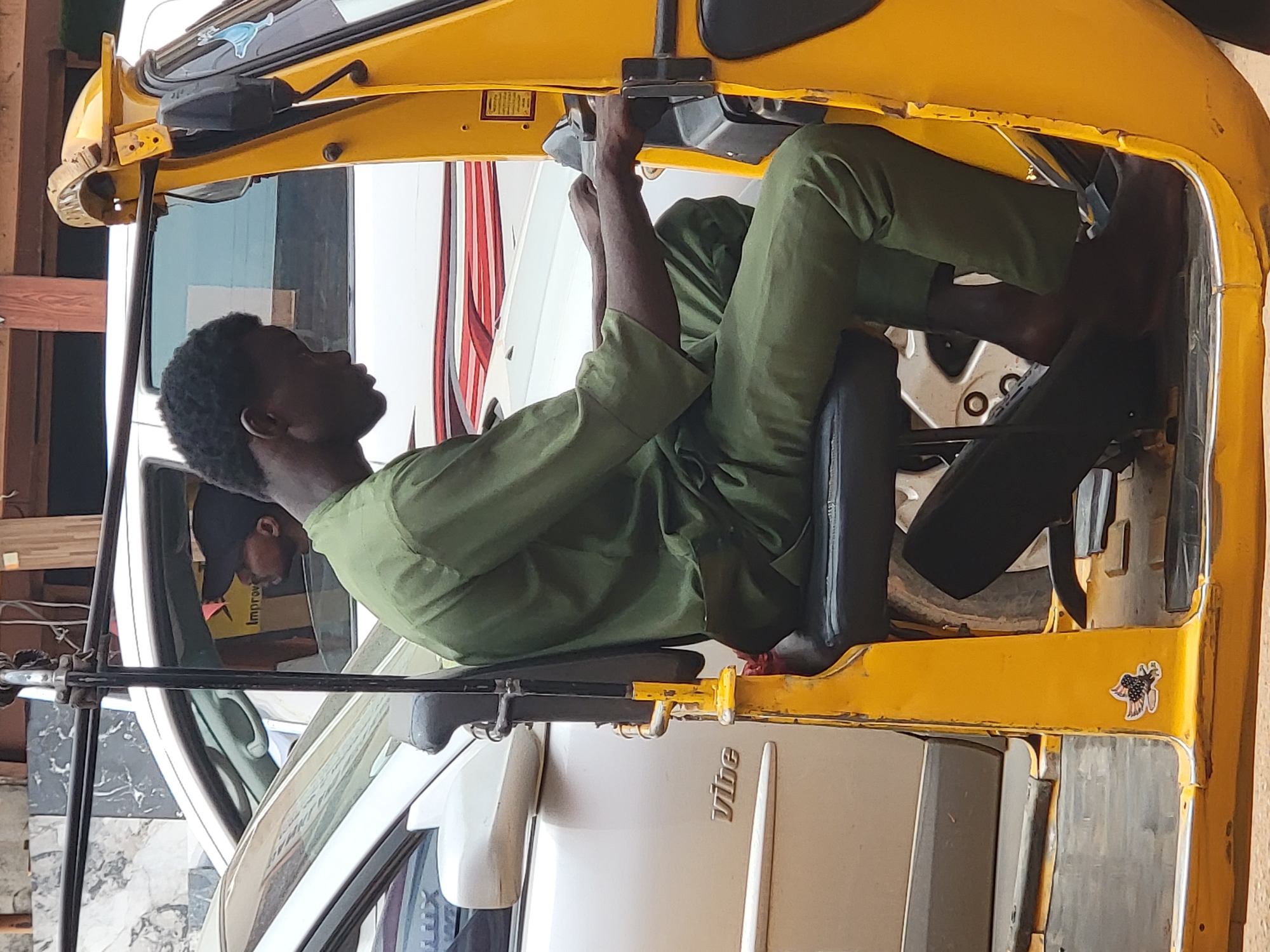 A man is working on a yellow three-wheeled vehicle, identified as a Bajaj Qute, in Maiburiji, Nigeria. The man, wearing green clothing, is seated inside the vehicle and appears to be focused on a task near the dashboard. The Bajaj Qute is parked next to a silver car, partially visible in the frame. The scene suggests a repair or maintenance activity taking place, possibly in a garage or workshop setting.