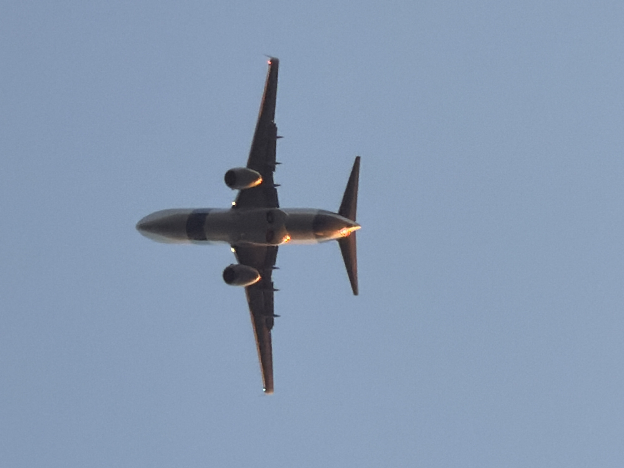 An airplane is captured in flight from a low-angle perspective, seen against a clear, pale blue sky. The jet aircraft, likely a commercial airliner given its size and engine configuration, is illuminated by the sun from below and slightly behind, casting a warm, golden glow on the underside of its fuselage, engine nacelles, and the leading edges of its wings and tail fin. This lighting suggests either sunrise or sunset. Two engines are visible, mounted beneath the wings. The aircraft displays a dark gray or black band around its mid-fuselage. The tail section features a prominent vertical stabilizer. No clouds are visible, indicating clear weather conditions. The activity is the airplane flying overhead. No people are visible, nor is any visible text present on the aircraft or in the sky. The image context is Tel-Aviv, Israel.