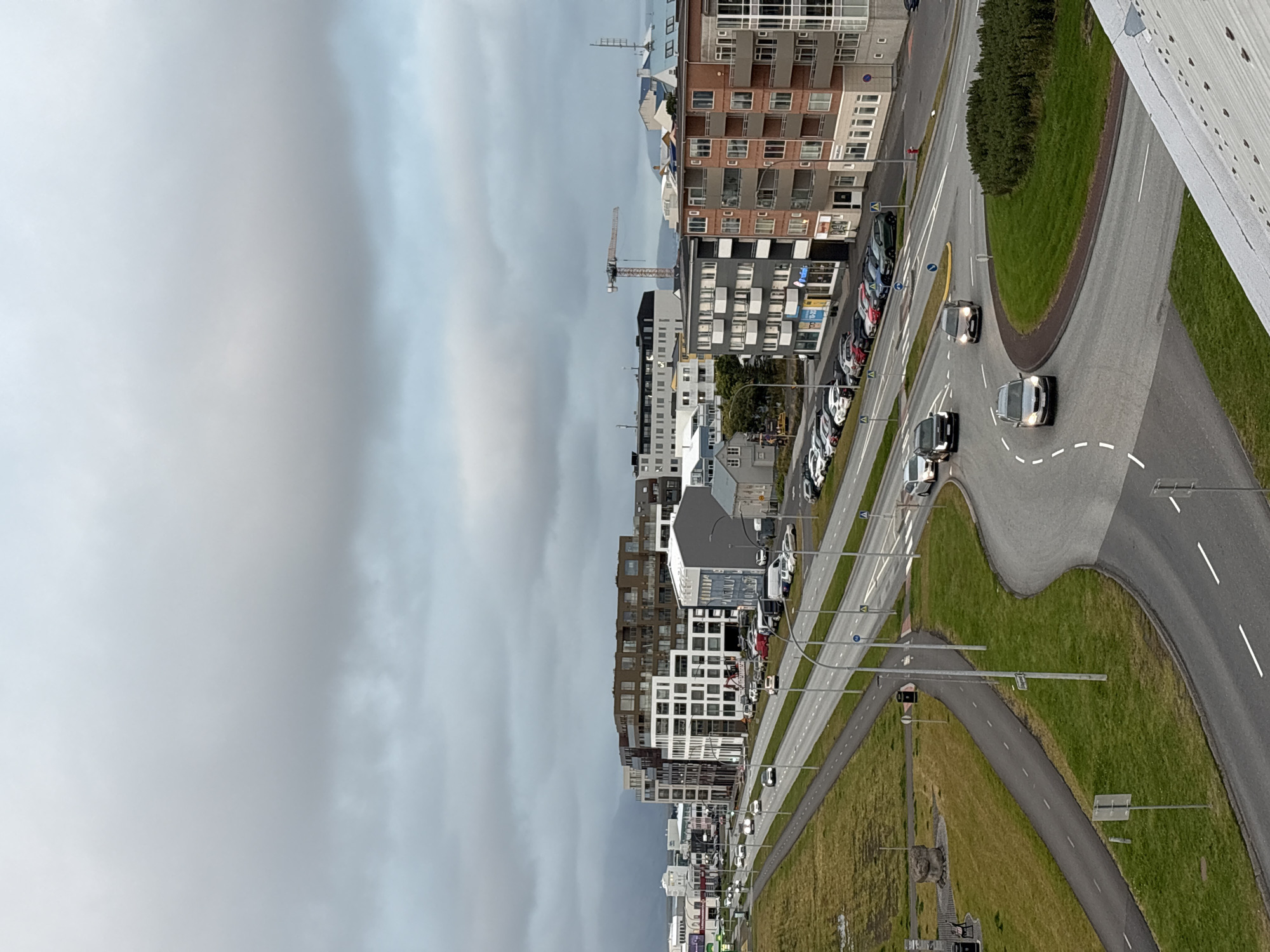 The image depicts an overcast day in Reykjavik, Iceland, with a view of a modern urban landscape dominated by multi-story buildings and a city street. In the foreground, a curved road network is visible with several cars in motion. One car, a silver Skoda, is turning, while a dark SUV and a grey car are also driving on the road. The road is lined with white dashed lines and there are pedestrian walkways and green spaces.
Further back, a large, contemporary building with a facade of white and dark brown panels stands prominently on the left side of the image. Adjacent to it are other buildings of varying architectural styles and colors, some featuring balconies. A construction crane is visible in the distance, suggesting ongoing development. A wider road with multiple lanes and streetlights runs parallel to the buildings, with more cars and parked vehicles on the side. The overall environment appears to be a commercial or residential area with a mix of new and older structures. The sky is heavily clouded, creating a subdued lighting condition.