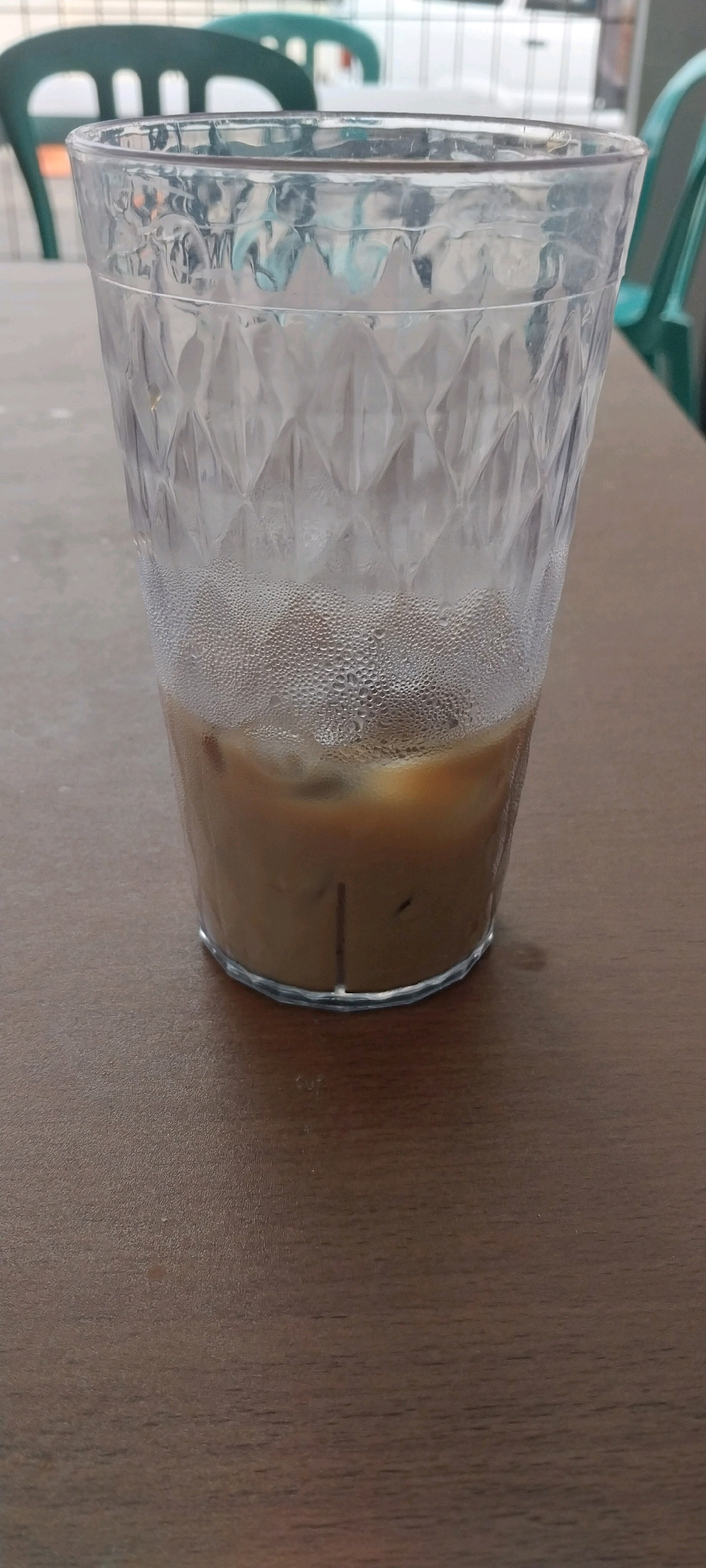 A tall, clear, textured glass filled with a light brown beverage, likely iced coffee, sits on a dark brown wooden table. The glass is partially full, with ice cubes visible beneath the liquid. The exterior of the glass is covered in condensation. In the blurred background, several green plastic chairs are visible, suggesting an outdoor seating area, possibly at a cafe or restaurant. The scene appears to be a close-up shot focused on the drink, with the background elements out of focus, indicating a shallow depth of field. The overall lighting suggests an ambient daylight setting. The implied location in South Tangerang, Indonesia, suggests a warm climate where cold beverages like iced coffee are commonly consumed.