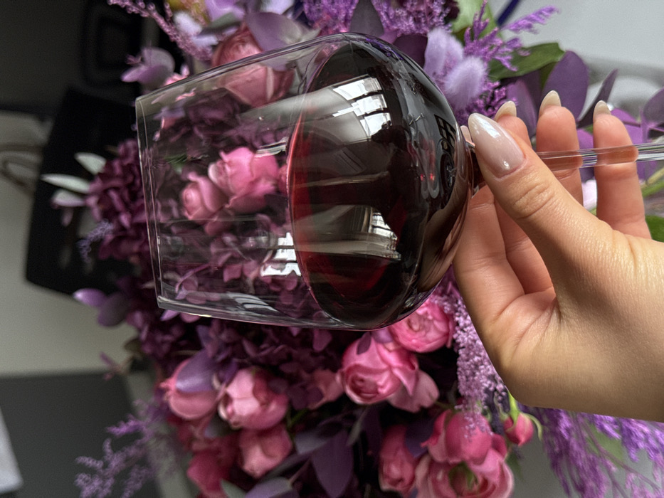A close-up, high-angle photograph captures a fair-skinned hand holding a clear, stemmed wine glass containing dark red wine. The hand exhibits manicured nails with a light pink or nude polish. Reflections visible on the wine glass's surface suggest an indoor setting with vertical light sources, possibly windows or blinds. The background features a large, out-of-focus bouquet of flowers, predominantly composed of pink roses, purple flowers, and lavender foliage. A dark, indistinct object is visible behind the floral arrangement. The scene is located indoors in the city of Yerevan, Armenia.