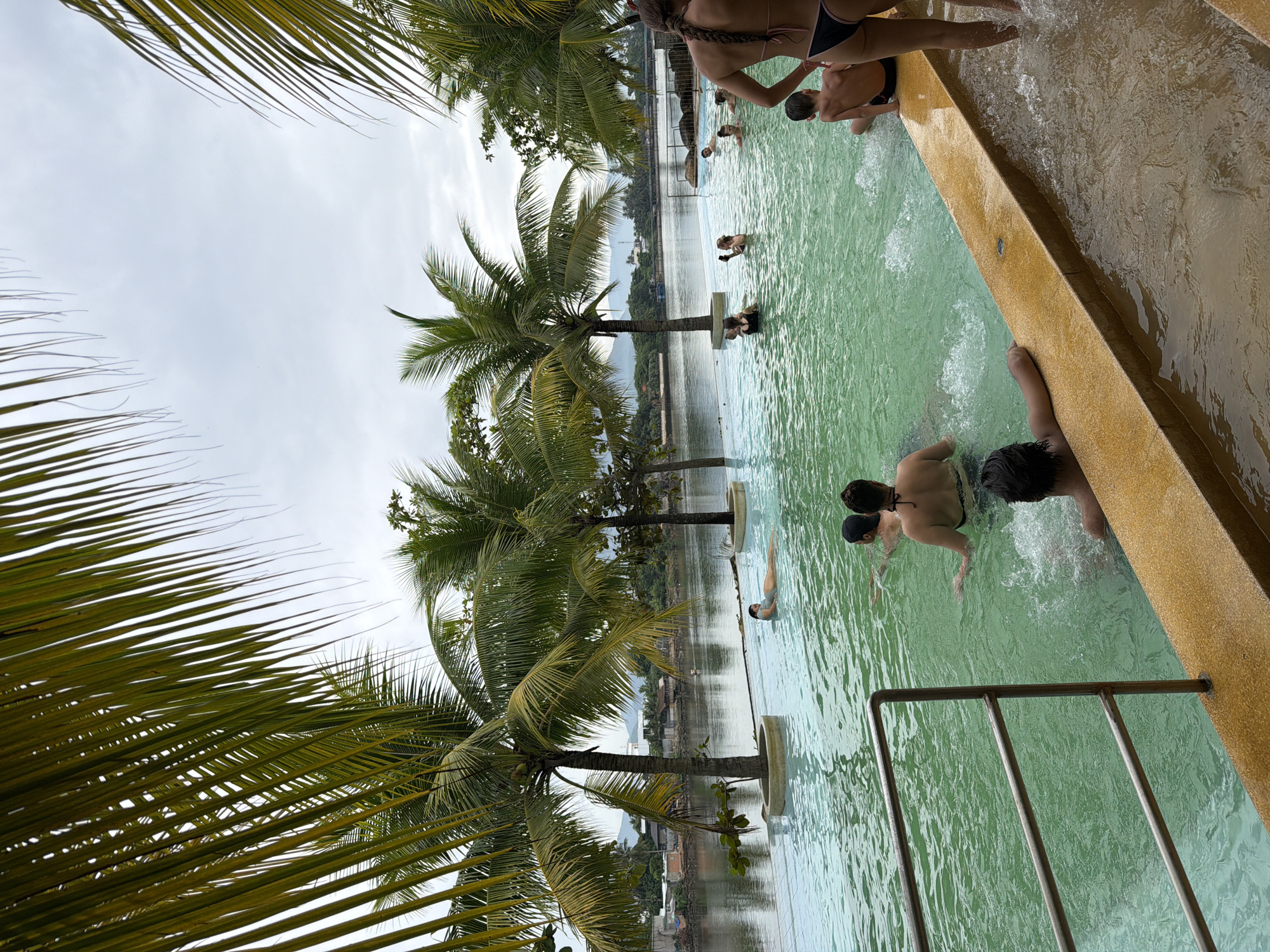 The image depicts a swimming pool at the Vinpearl Hotel Nha Trang, located on Hon Tre Island in Nha Trang, Vietnam. The pool features clear, turquoise water and is surrounded by lush palm trees. Several people are enjoying the water, including a woman floating on her back, a group of individuals swimming and splashing, and children playing. In the background, the Nha Trang coastline is visible, with its characteristic mountainous terrain and urban development. The sky is overcast, suggesting a humid tropical climate. The scene is one of relaxation and recreation, with a focus on the natural beauty of the surroundings and the enjoyment of the guests.