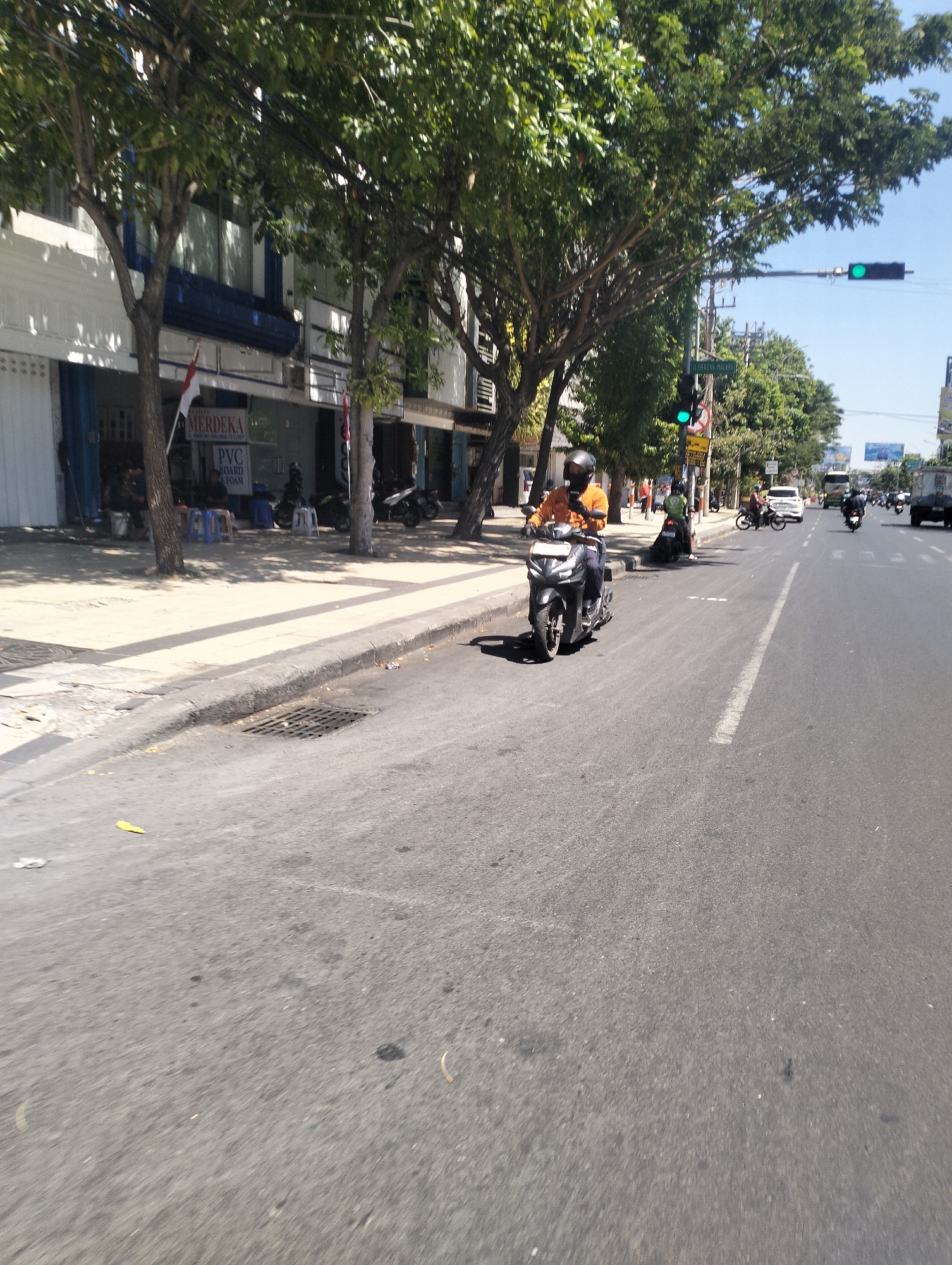 This image depicts a street scene in Surabaya, Indonesia, on a sunny day. In the foreground, a dark-colored motorcycle is traveling on a paved road. The rider, wearing an orange shirt and a black helmet, is the central subject. Behind the motorcycle, other vehicles, including cars and motorcycles, are also visible on the road, moving in the same direction.
To the left of the road, a sidewalk runs alongside a row of buildings. The buildings have a white facade with blue accents and feature storefronts. Trees with lush green foliage line the sidewalk, providing shade. A street sign and a traffic light are visible further down the road. The sky is clear and blue. The overall impression is a typical urban street with moderate traffic and pedestrians.