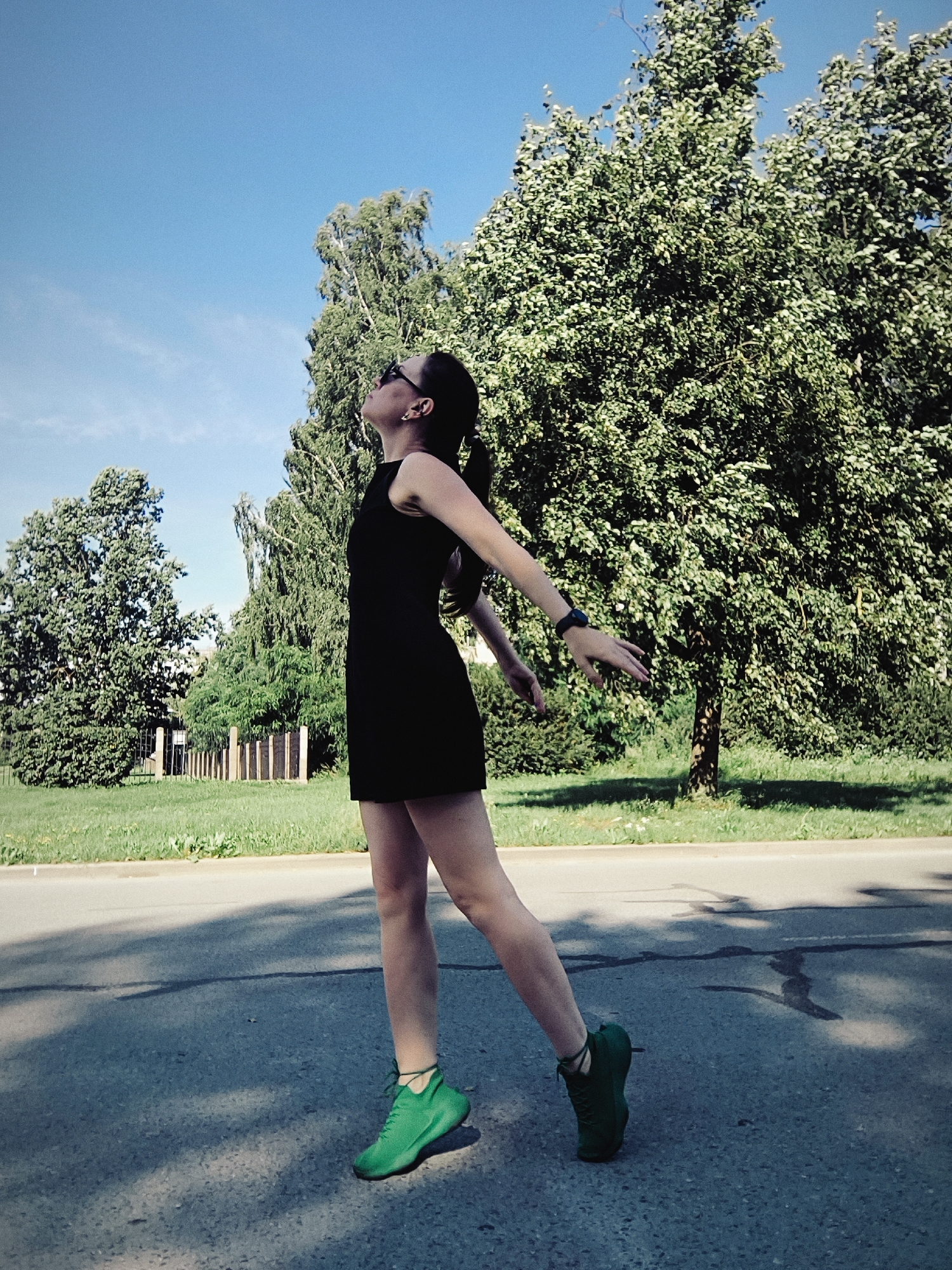 A young woman in a black dress and bright green sneakers stands on an asphalt road, her head tilted back and arms outstretched, looking up at the sky. She has sunglasses on and her hair is pulled back in a ponytail. The background features a lush green park with trees and a clear blue sky. This scene is likely located in Riga, Latvia, given the visual characteristics of the trees, greenery, and the overall park-like environment often found in the city's green spaces.