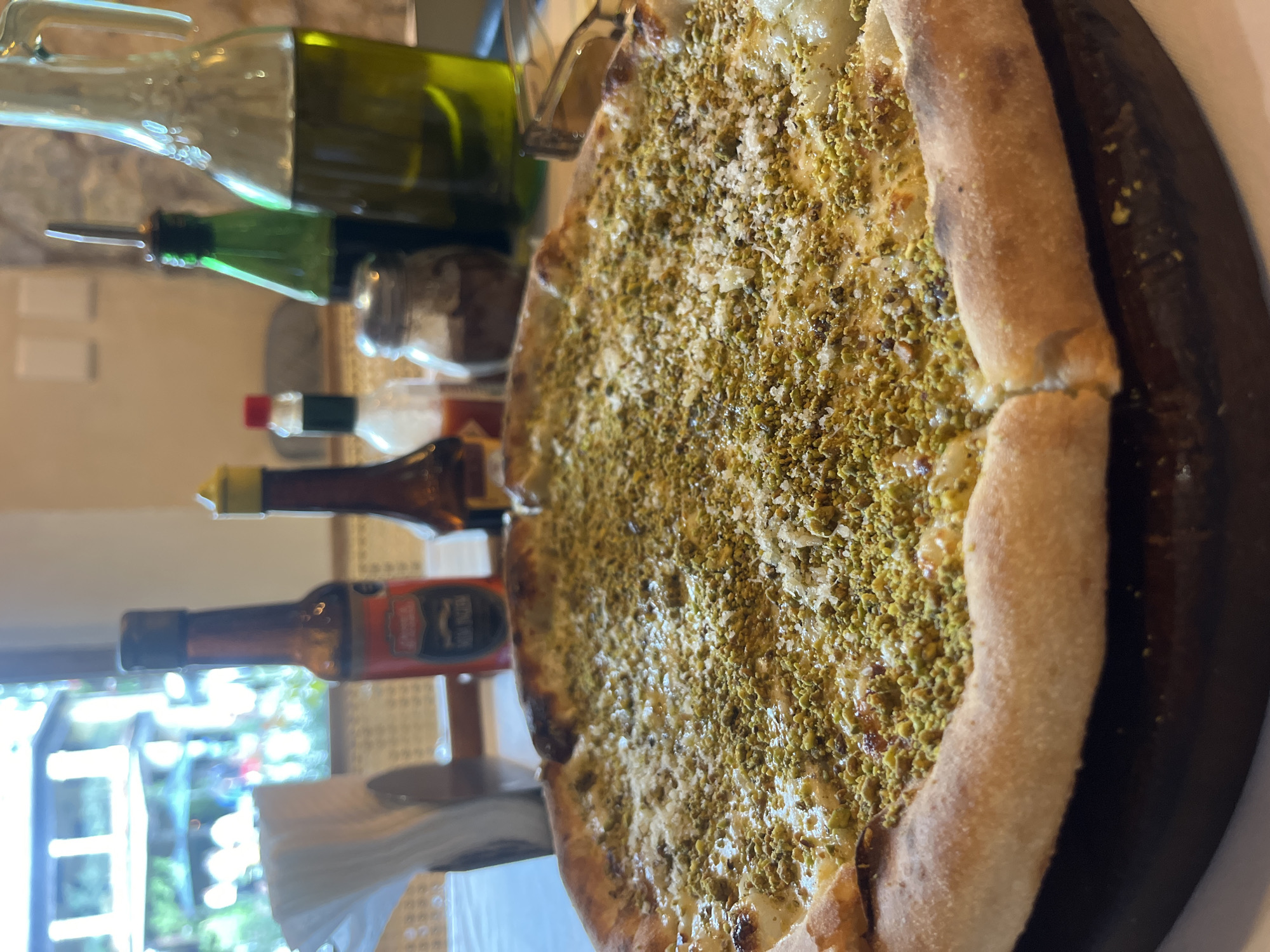 A high-angle, close-up shot features a pizza topped with what appears to be pistachio and shredded cheese. The pizza is placed on a dark-colored wooden board. Behind the pizza, various bottles are visible, including oil and sauce bottles. The scene is positioned indoors, near a window offering a glimpse of the outside environment, which seems to be a garden or street. The location in the city of Domingo Arenas, Mexico, is not directly identifiable from the image.