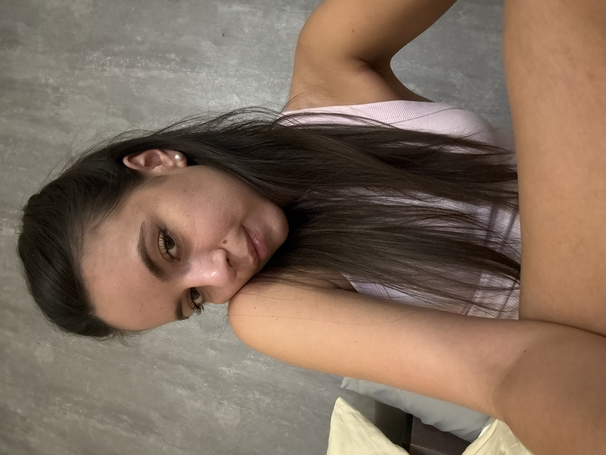 This media file shows a close-up, rotated portrait of a young woman. She has long, dark brown hair, fair skin, and brown eyes. She is wearing a light pink top and a pearl earring. Her head is tilted, and her right arm is bent, with her hand resting near her face. Her expression appears relaxed and contemplative. The background is a textured gray wall, suggesting an indoor setting. The lighting is soft, indicating it could be daytime or evening with indoor illumination. No text is visible in the image.