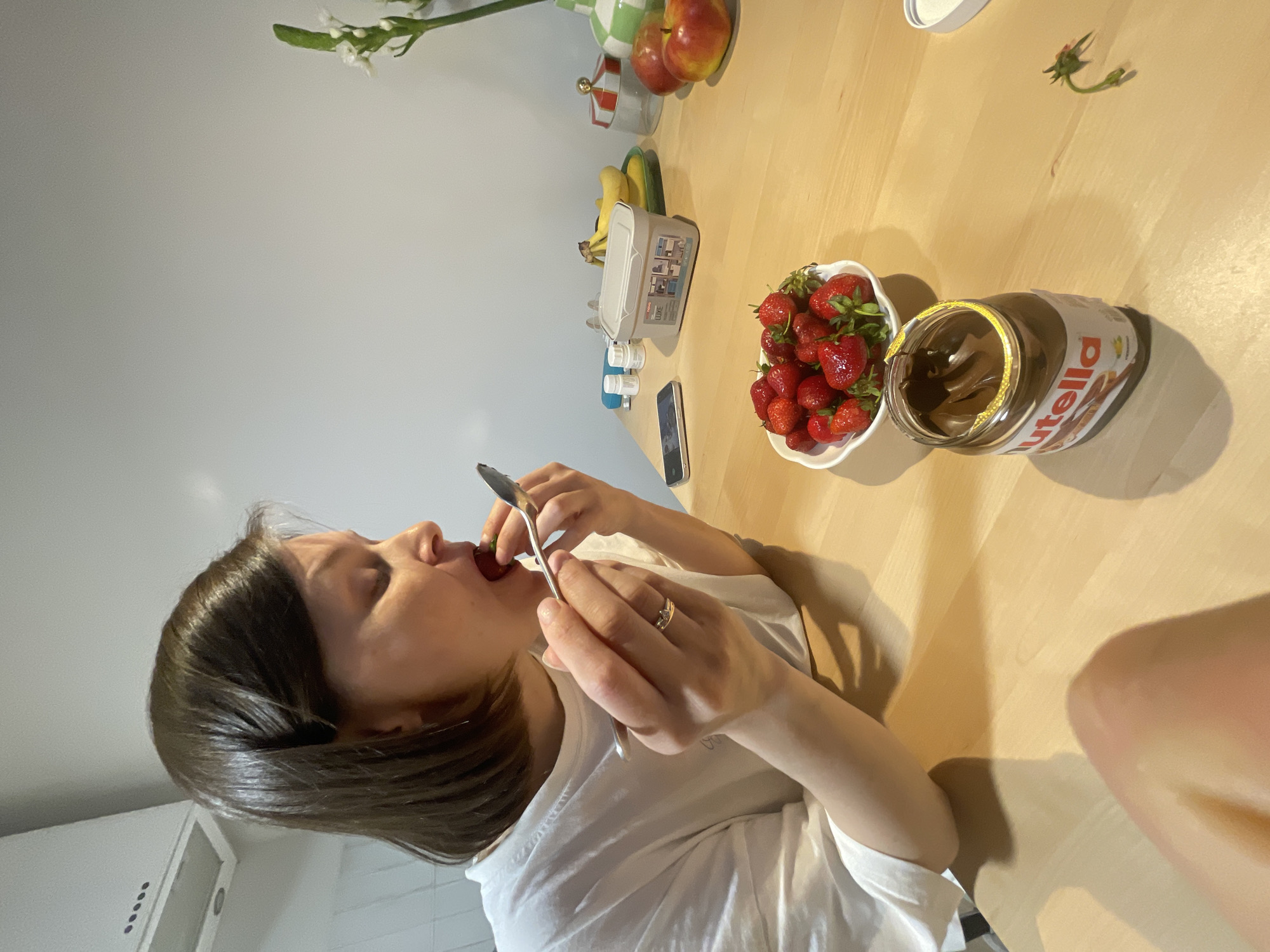 A woman with dark hair wearing a white t-shirt is sitting at a wooden table and eating a strawberry with a spoon. On the table are a bowl of strawberries, a jar of Nutella, a smartphone, a white container, bananas, apples, and a vase of flowers. The setting appears to be a kitchen. There is no information available to determine if this image was taken in Izhevsk, Russia.