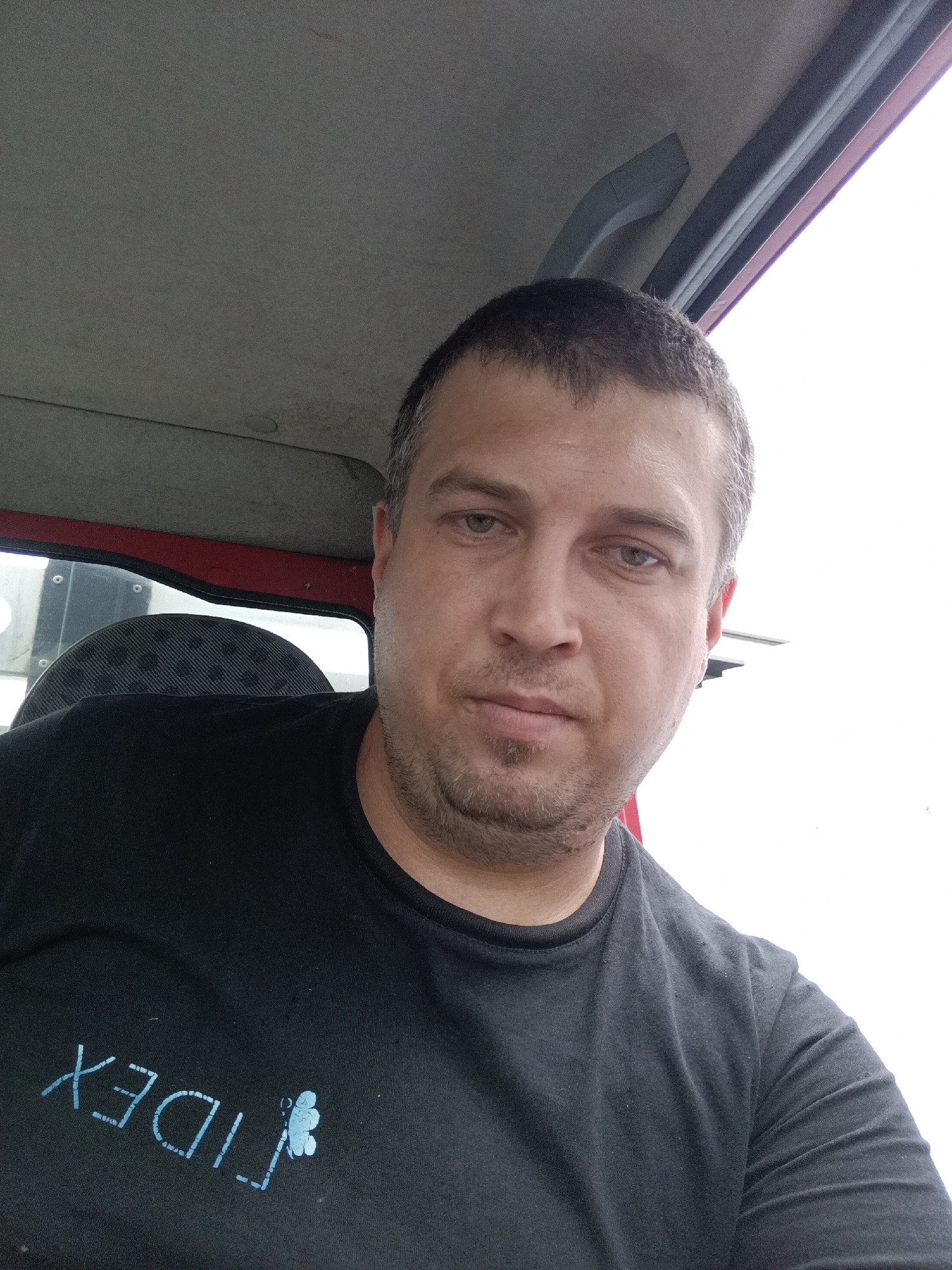 In Svémyslice, Czechia, a man is taking a selfie inside a tractor cabin. The man, appearing to be in his late 30s or early 40s, has short, dark hair with some graying at the temples and light stubble. He is wearing a dark t-shirt with a blue logo that appears to read "FIDEX" in a stylized font, accompanied by a small, light blue graphic of three dots arranged in a clover-like shape. The cabin's interior is visible, featuring a gray headliner, a red frame around the window, and a dark, textured seat. The exterior light suggests it is daytime.