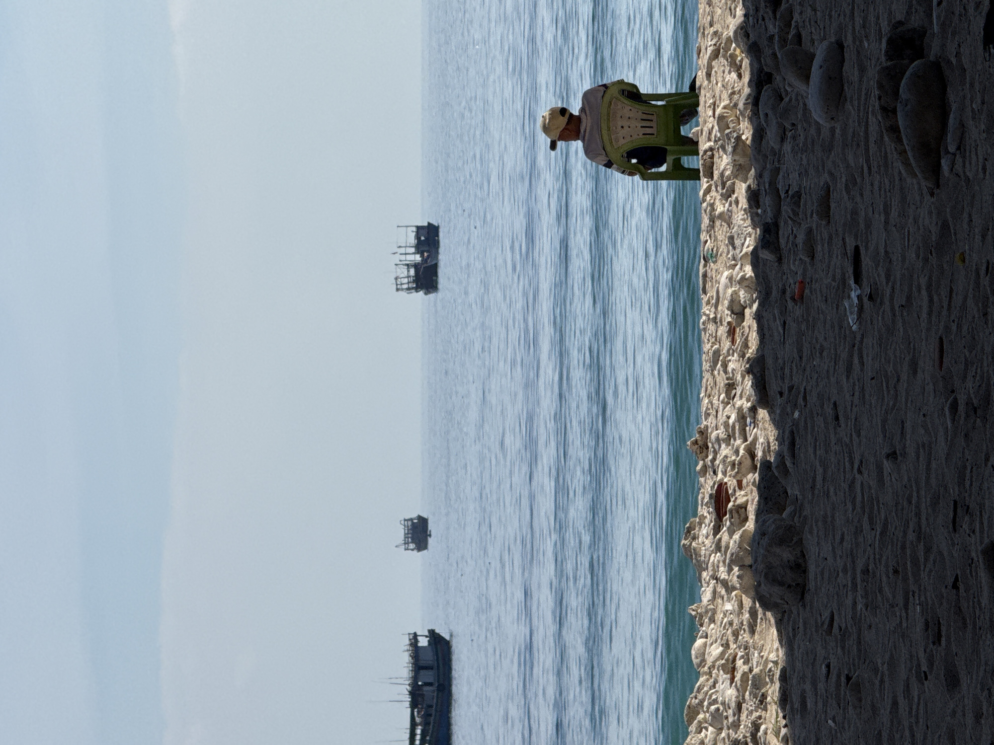 A man wearing a hat and a striped shirt sits on a green plastic chair on a beach. He faces the ocean, where several boats are visible in the distance. The beach is covered with sand and rocks, and the ocean is calm with gentle waves. The sky is clear and blue. The scene appears to be a serene, coastal landscape on Lý Sơn Island, Vietnam.
