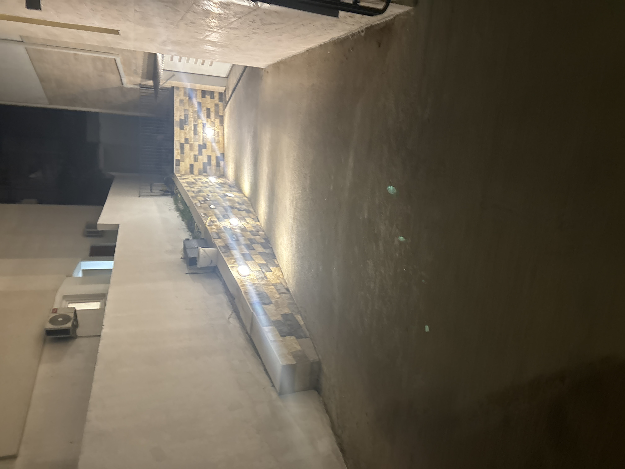 The image shows an outdoor paved area at night, illuminated by artificial lighting. A wall with decorative stone tiling runs along one side, with lights embedded in it, casting light onto the ground. To the left, a concrete wall extends upwards, and an air conditioning unit is visible mounted on it. A small raised concrete seating area is present in the foreground on the left. The ground is paved with a rough, grey material. In the background, there are buildings and a dark, unlit area suggesting it is nighttime. There are no people or visible actions in the image.