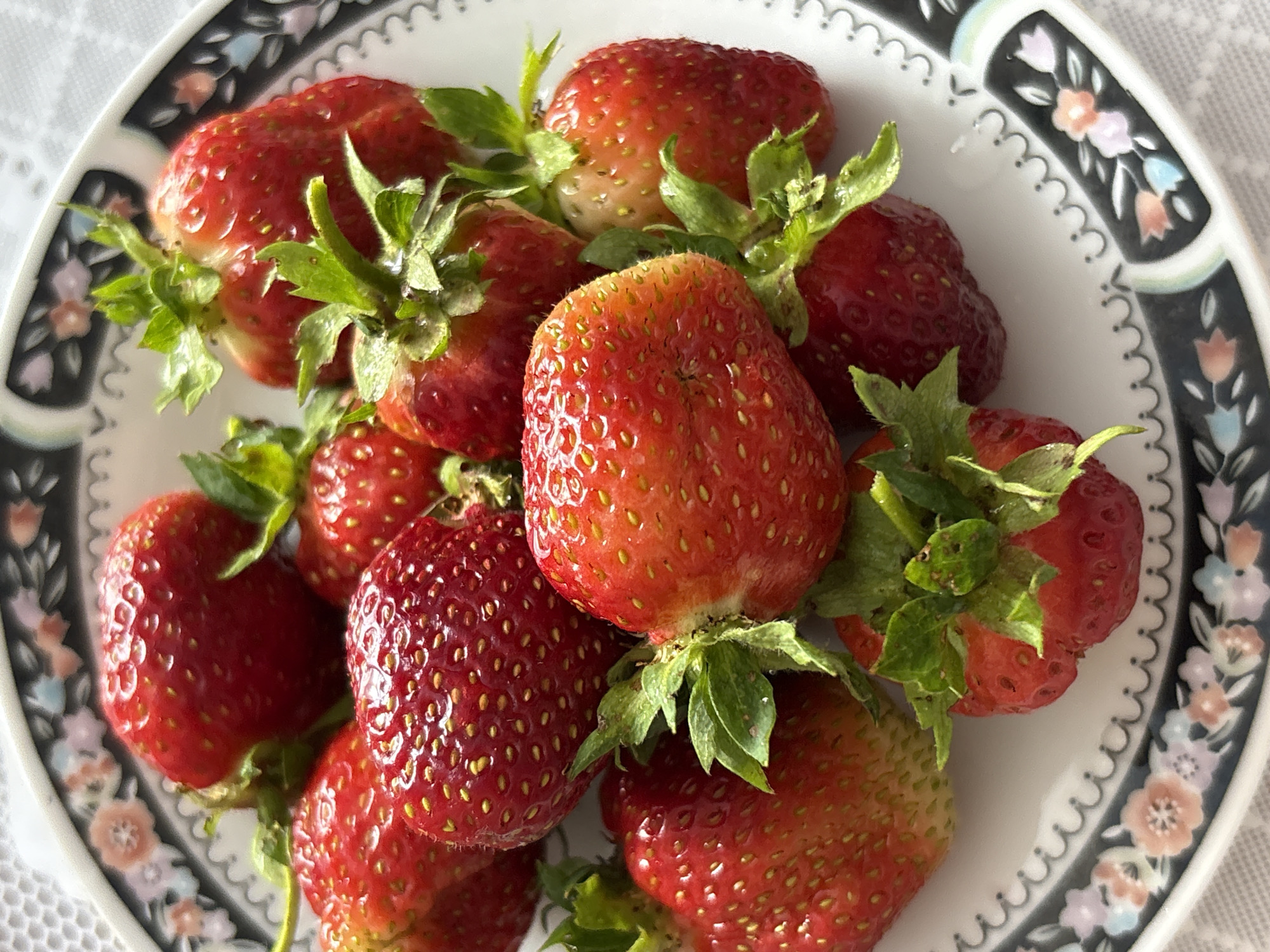 A close-up, overhead shot shows a white plate with a decorative floral rim filled with ripe strawberries. The strawberries are various sizes, predominantly red with some yellow seeds visible. Green stems and leaves are attached to some of the fruits. The plate rests on a white textured surface, possibly a tablecloth. The image does not contain information about the city of Kumyony, Russia.