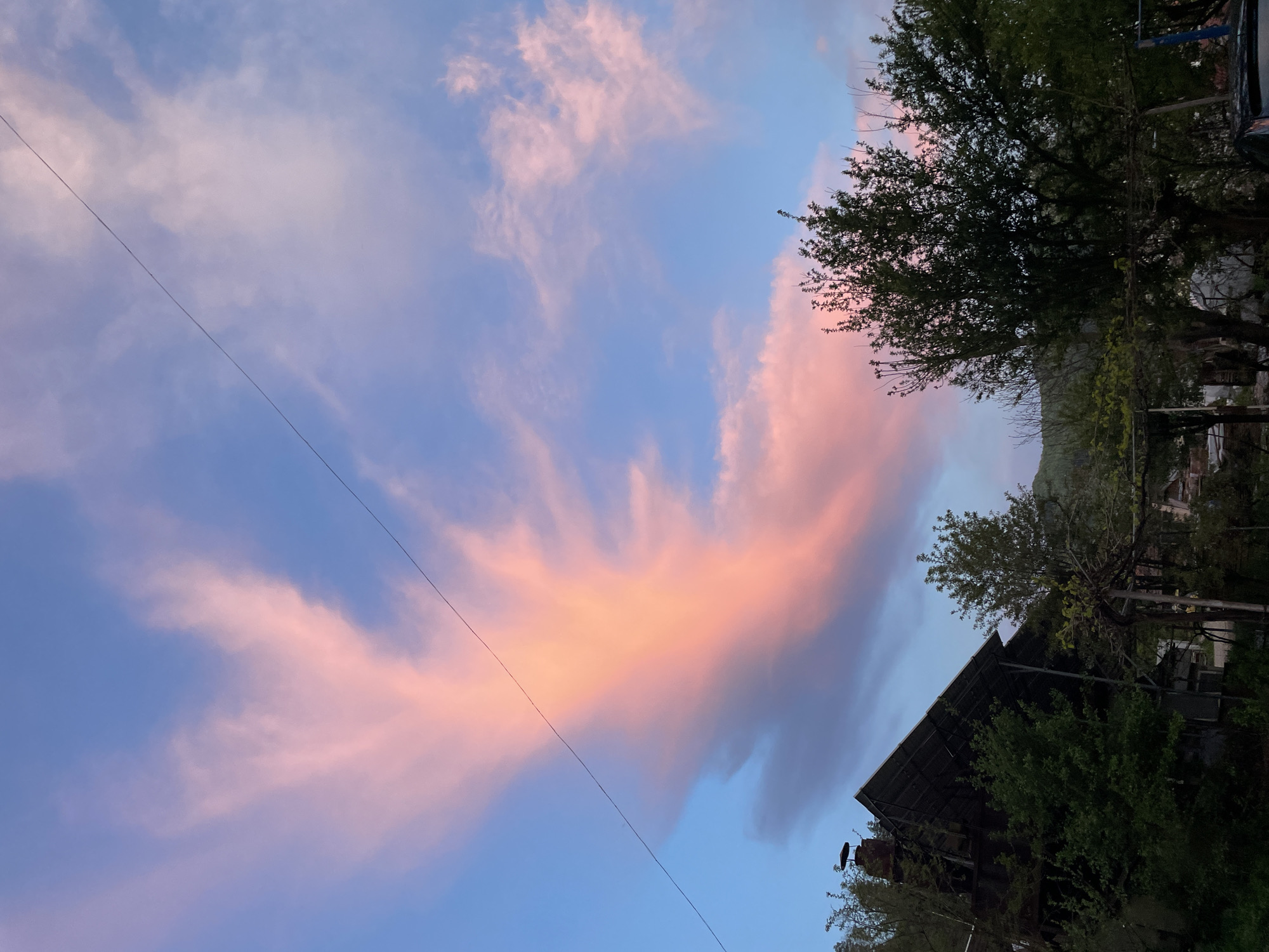 The image shows a sky with pink-tinged clouds and areas of blue. A power line stretches diagonally across the frame. The edge of a building with a dark roof is visible in the lower right. Green trees surround the building and are also present at the top of the image. The location is Zlatograd, Bulgaria.