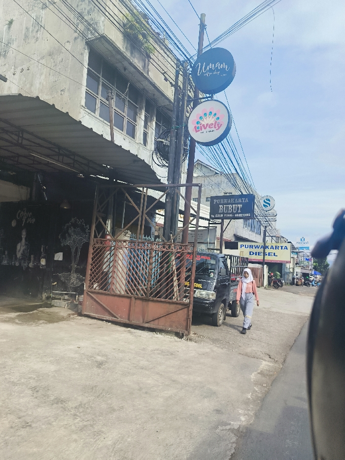 The scene is a street in Purwakarta, Indonesia, captured from a vehicle. The location is determined by the coordinates provided: latitude -6.543, longitude 107.441. On the left side of the street, there is a building with a sign that reads "Umami Coffee Shop". The building also has a large, red metal gate that is closed. On the opposite side of the street, a woman in a white hijab and a blue and white patterned dress is walking away from the camera. There is a white truck parked on the side of the street with "PURWAKARTA BURUT" written on it. Behind the truck is a sign for "PURWAKARTA DIESEL". On the street, there are electrical wires hanging overhead. There are also several other buildings in the background, including a building with the sign "Lively". The scene is captured on a bright, sunny day. The street is paved with concrete. The overall tone of the scene is mundane and ordinary.