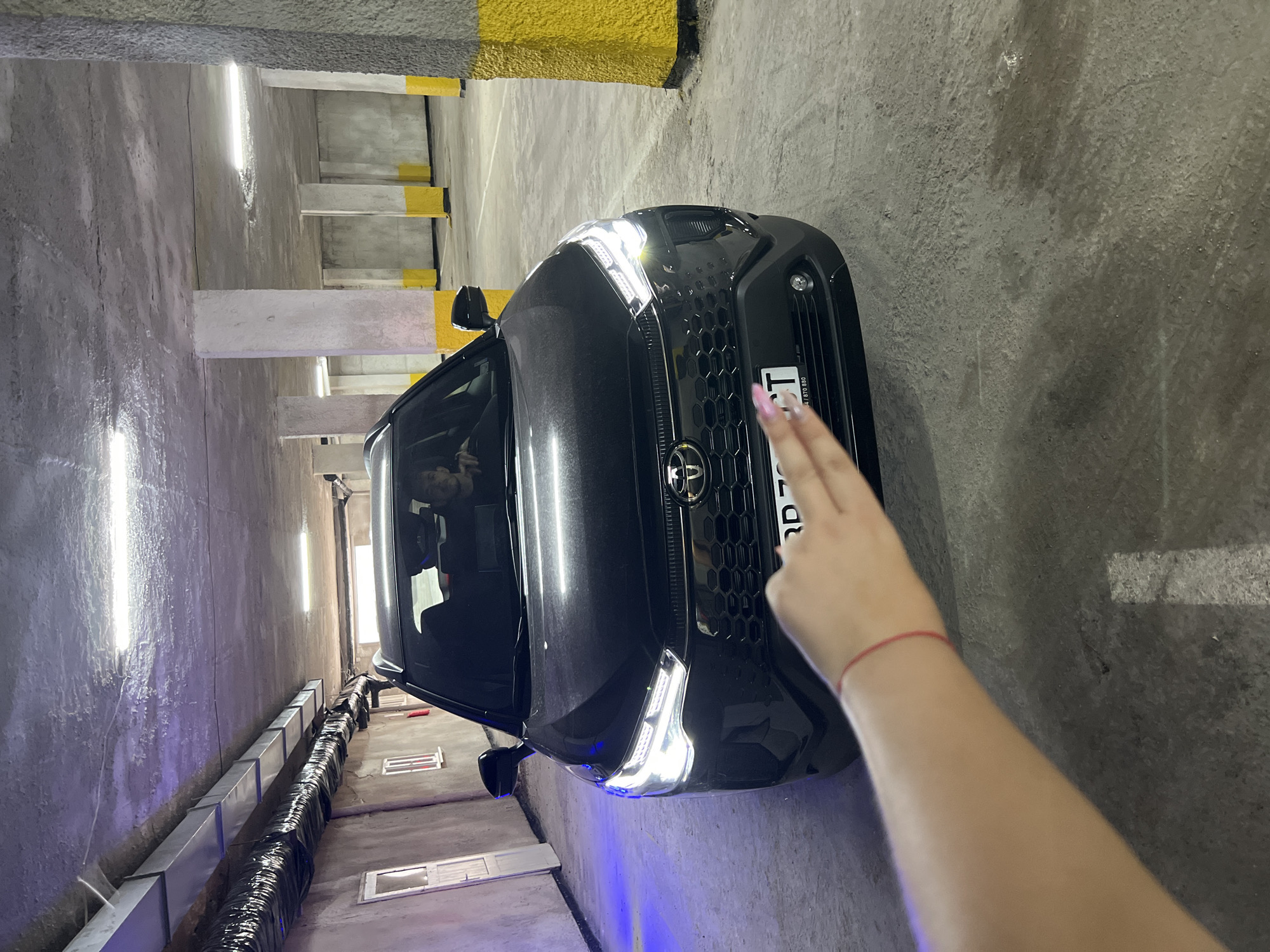 The image depicts a dark-colored Toyota RAV4 parked in an indoor parking garage. The car's headlights are illuminated. A person's arm and hand, with manicured fingernails, are visible in the foreground, obscuring the lower portion of the car's grille and license plate. The garage environment features concrete columns painted yellow at the base and a textured concrete ceiling with exposed ductwork and lighting fixtures. The floor is also concrete. The overall lighting is dim, with artificial light sources providing illumination. The location is identified as Vratsa, Bulgaria.
