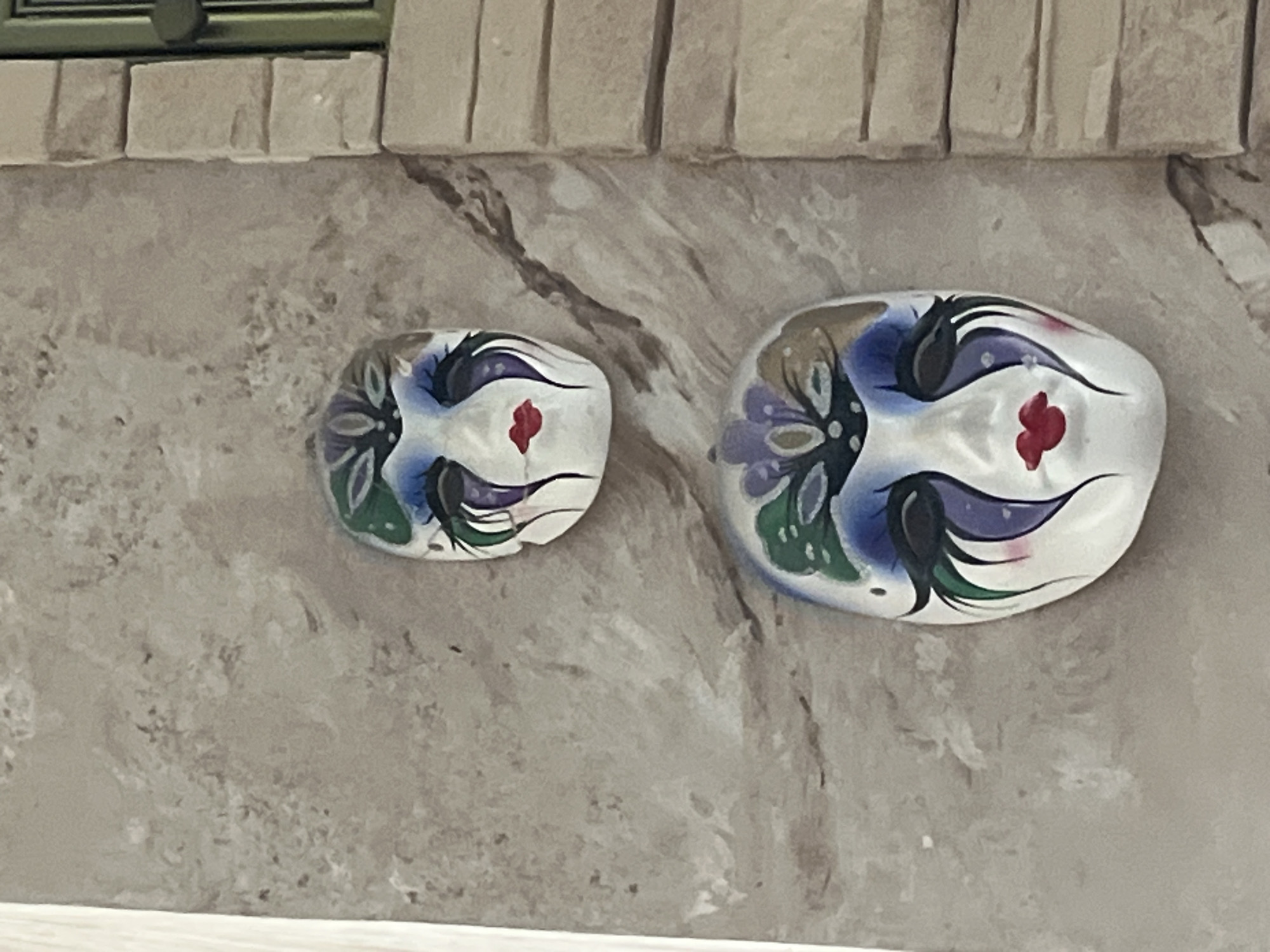 Two decorative masks are affixed to a textured, neutral-colored wall. The masks are identical in design, featuring painted details of exaggerated eyelashes, blue and purple eye makeup, and red lips. A floral-like pattern in shades of purple and green adorns the forehead area of each mask. The wall appears to be an interior or exterior surface with a painted or stucco finish. To the right of the wall, a section of light-colored brickwork is visible, suggesting a building facade or architectural feature. The image is taken from a slightly upward angle. There are no people or actions depicted in the scene. The location in Vladimirovka, Russia, is not visually evident from the image itself.