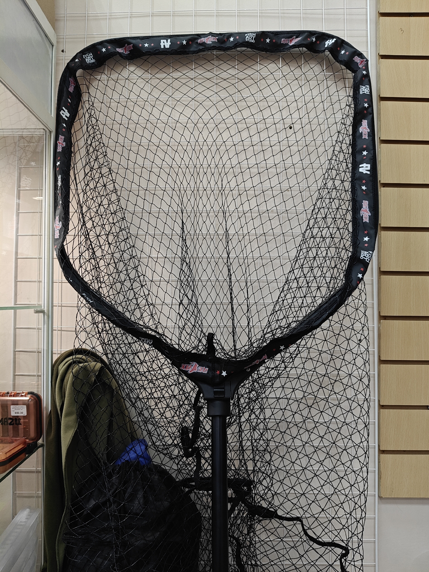 A fishing net hangs on a wire grid in a store. The net has a black frame with red and white stars and the text "FISH ART STUDIO" and "REFOX". A green jacket and a black bag are draped behind the net. A display case with a box labeled "MAZU" is visible to the left. The store's location is Chaikovsky, Russia. Wood paneling is visible to the right of the net.