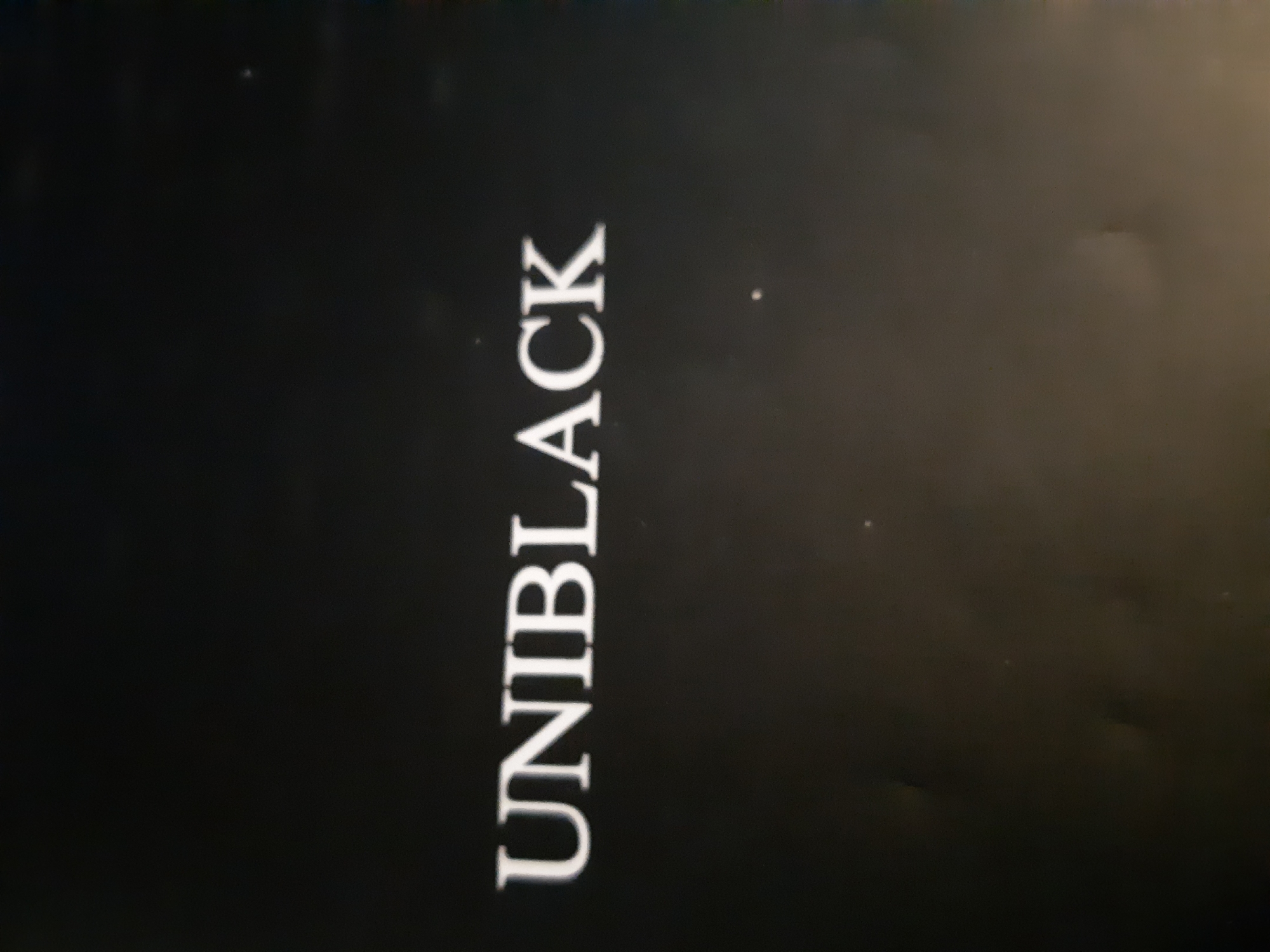 The image shows the word "UNIBLACK" in white, vertical text against a dark, possibly black, background. The background is not uniformly dark, with variations in shading and some minor imperfections or dust specks visible. The photo appears to be slightly out of focus. There is no indication of the location being Plovdiv, Bulgaria, or any people or other subjects.