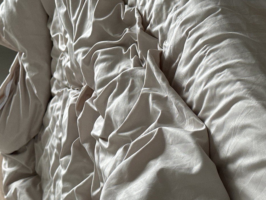 The image displays an unmade bed, fully covered by light-colored, possibly beige or pale taupe, bedding. The fabric exhibits extensive rumpling, folding, and creasing across its surface. Light reflects off the material, indicating a smooth or slightly lustrous texture. Shadows are present within the folds, suggesting an overhead or diffuse light source. No individuals, identifiable objects beyond the bedding, or specific actions are visible. The environment is an interior space, consistent with a bedroom. The precise location within the city of Yekaterinburg, Russia, cannot be determined from the visual evidence presented in the image.