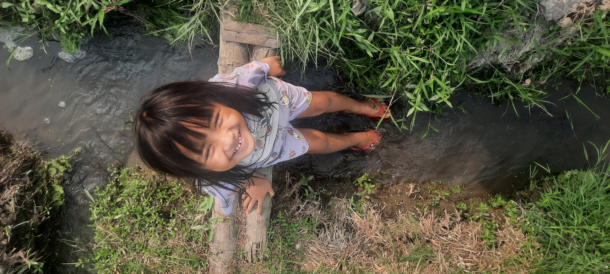 This media file shows a young girl with dark hair, wearing a light purple t-shirt with a cartoon print, and red flip-flops. She is sitting on a wooden plank laid across a narrow, shallow stream. Her legs are submerged in the water, and she is smiling broadly with her eyes closed, appearing joyful. The stream is surrounded by lush green grass and vegetation. The overall scene suggests a warm, sunny day, likely in a rural or natural setting, possibly during daylight hours given the lighting. The image evokes a sense of childhood innocence and simple pleasure. The location is identified as Ploso, Indonesia, which aligns with the tropical-looking vegetation.