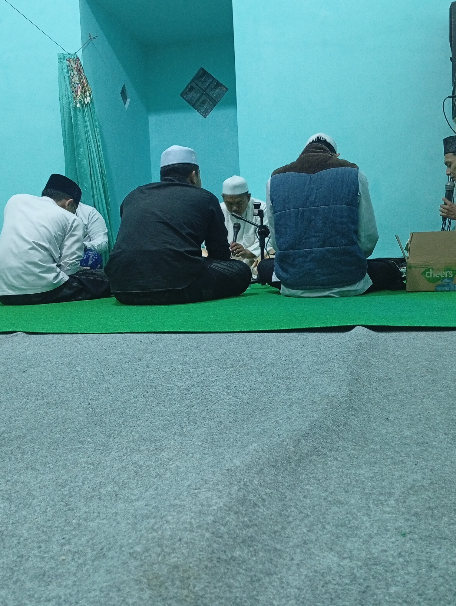 Four men are seated on a green carpet in what appears to be a religious gathering. The man on the far left is wearing a white shirt and a black cap. Next to him, a man in a black shirt and cap is facing away from the camera. In the center, a man in a white cap and shirt is speaking into a microphone. To his right, a man wearing a blue vest over a white shirt and a white cap is also facing away from the camera. A cardboard box with the word "cheers" is visible on the right side of the image. The walls are painted a light blue, and a decorative cloth hangs on the left. The scene suggests a religious sermon or prayer session taking place indoors, possibly in Jatisari, Indonesia, given the context.