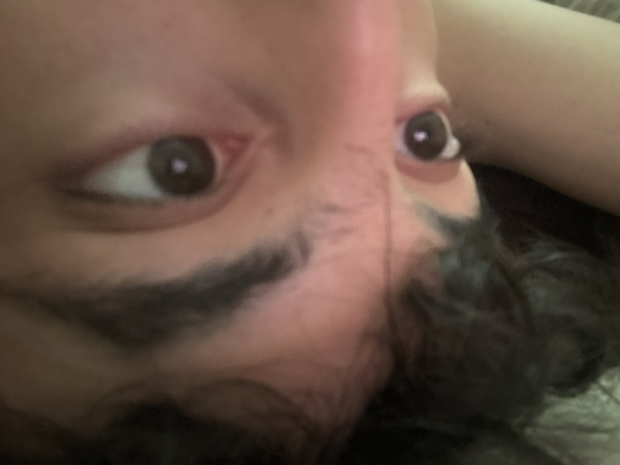 The image is a close-up, vertically oriented, portrait-style photograph of a person's face. The perspective is extreme, with the camera positioned very close to the subject, resulting in a fragmented and somewhat abstract view of their features. The subject appears to be looking directly at the camera, with their eyes being a prominent focal point. The lighting is dim and appears to be coming from above and to the right of the subject, creating soft shadows and highlighting the contours of their face. The overall tone of the image is warm, with skin tones appearing reddish-pink and shadows being a deep brown. The image is out of focus in most areas, with only small sections, such as the eyes and parts of the skin, being relatively sharper. The presence of dark, curly hair frames the face on the left side of the image. The background is largely indistinct and appears to be a continuation of the subject's skin or clothing, with a smooth, blurred texture.