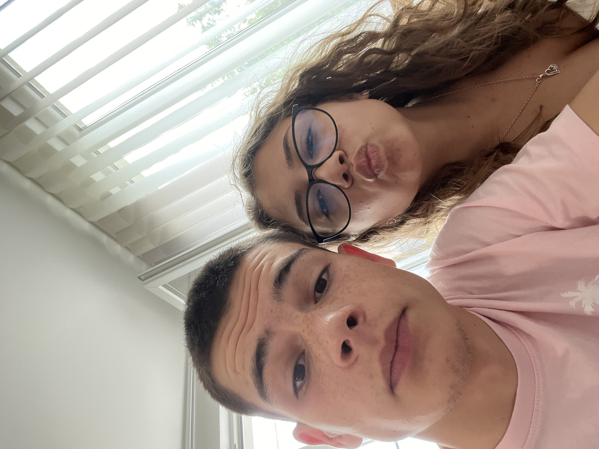 A selfie shows a young man and woman positioned close to each other. The man is in the foreground, and the woman is behind him, slightly angled. The man is wearing a pink t-shirt and has short dark hair. He has visible freckles. The woman is wearing glasses and is pursing her lips in a kissing expression. She has long, curly brown hair and is wearing a necklace with a heart pendant. In the background, there is a window with closed horizontal blinds, providing a source of light. The location is indoors. Karavelovo, Bulgaria, is not discernable from the image.